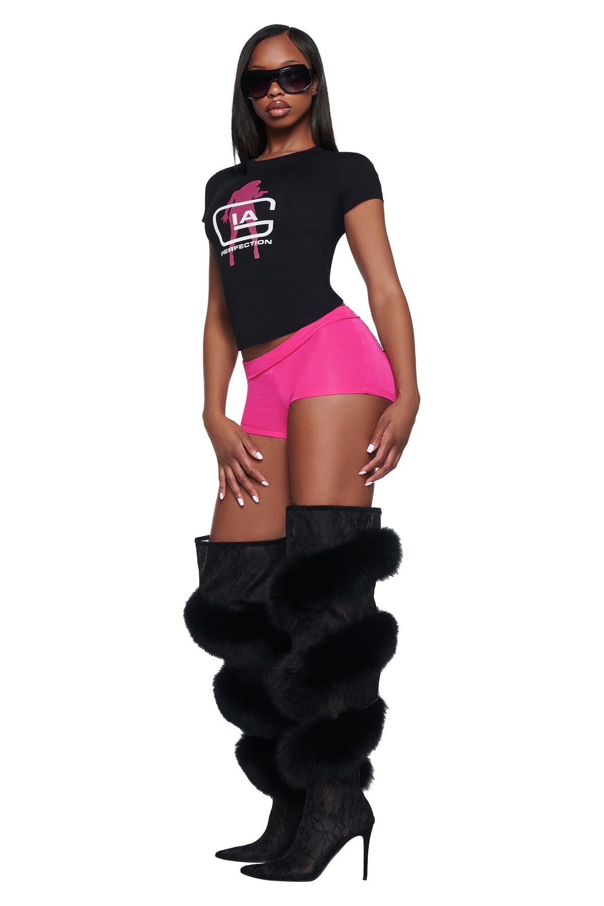 A person wearing a black I.AM.GIA YVL T-SHIRT - BLACK GIA cropped shirt with pink Young Vamp Life graphic text, hot pink shorts, thigh-high black boots with fur trim, and dark sunglasses.