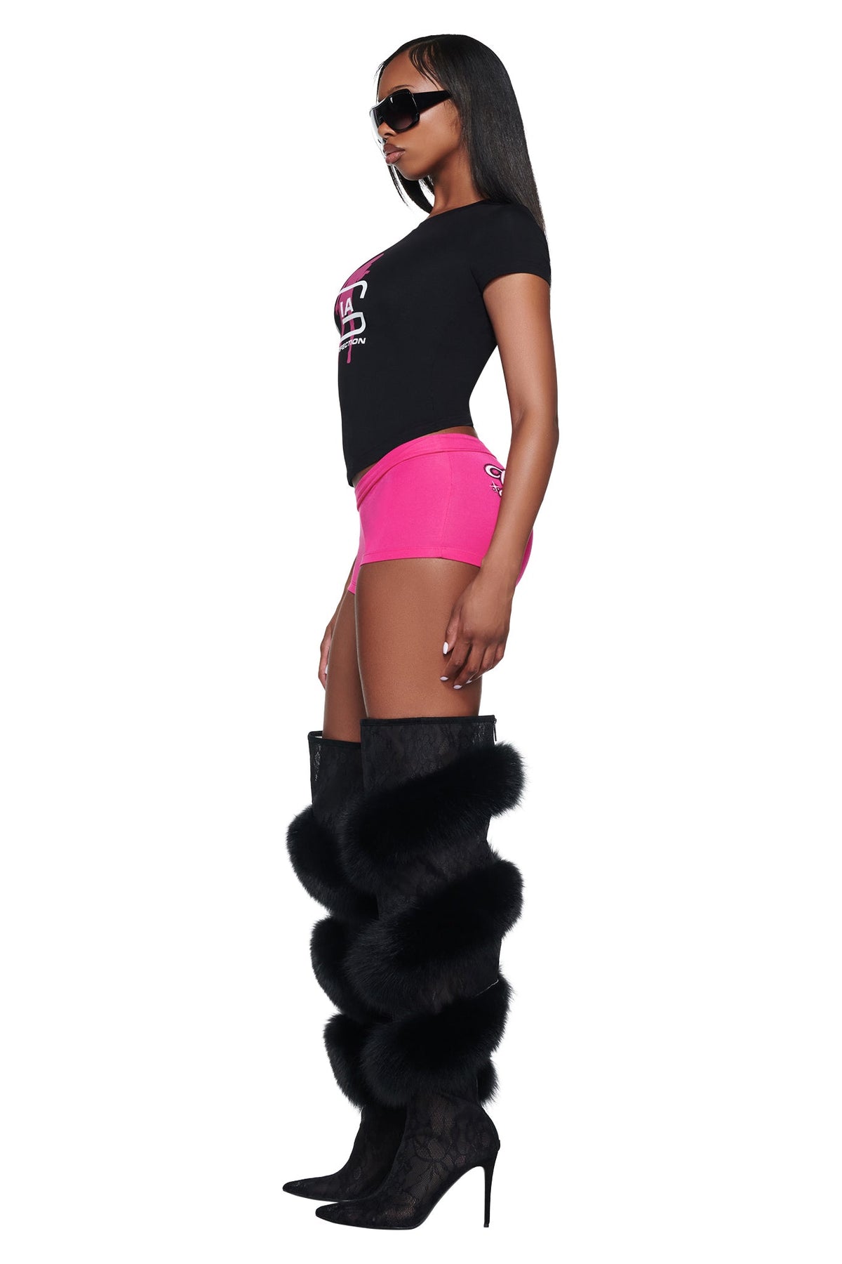 A model in profile wearing a black I.AM.GIA YVL T-SHIRT - BLACK GIA with a pink logo, pink shorts, and black fur-trimmed high-heeled boots.