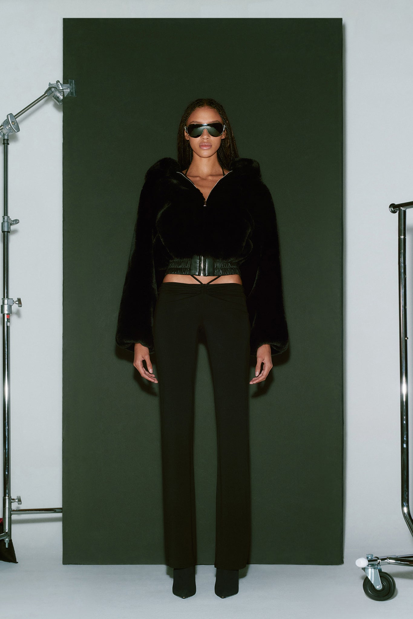 A woman is standing against a dark backdrop, dressed in the I.AM.GIA 717 JACKET - BLACK, high-waisted pants, and pointed shoes. She has long hair and wears large black sunglasses. The setting appears to be a photo studio with adjustable lighting equipment visible.