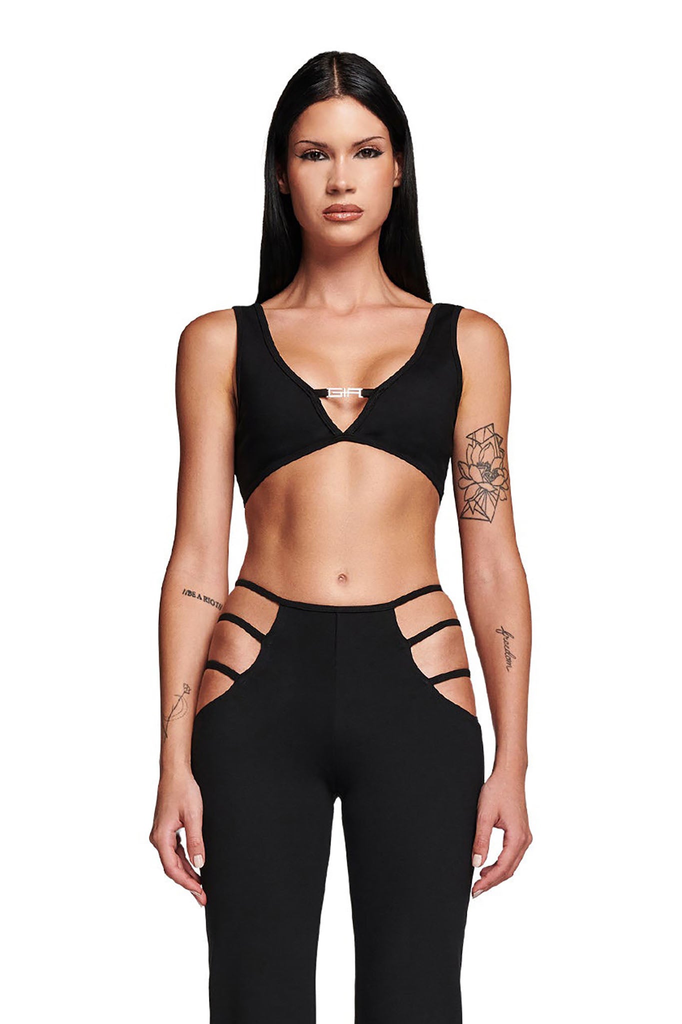 A person with long dark hair is wearing the black LUCID TOP by I.AM.GIA and bodycon fit black pants featuring side cutouts. They have tattoos on both arms and are standing against a plain white background.