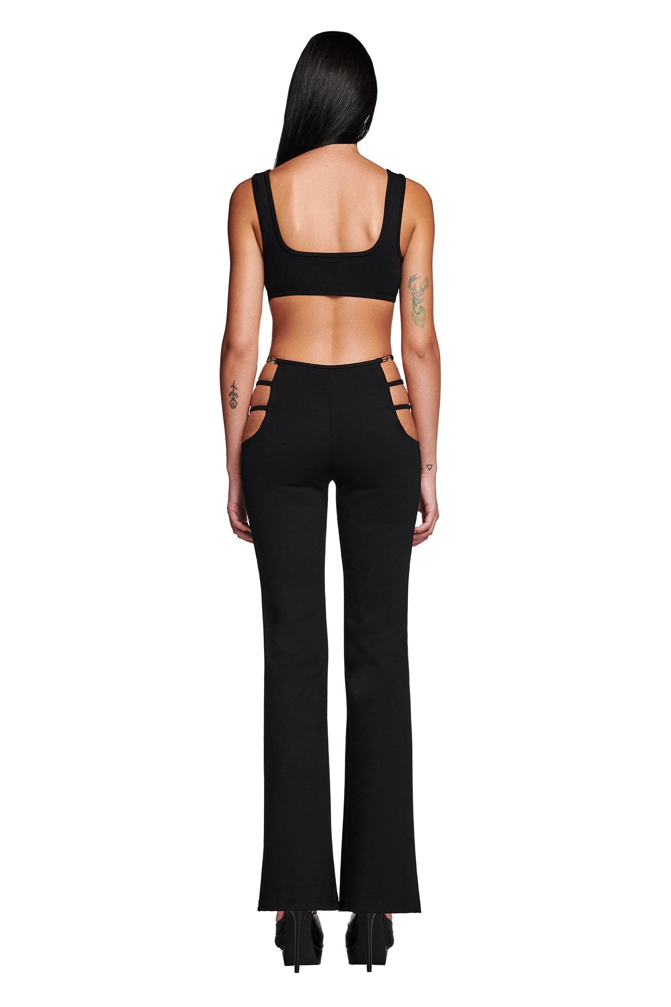 A woman with long dark hair wears a black LUCID TOP from I.AM.GIA, showcasing its bodycon fit. She pairs it with black high-waisted pants that have side cut-out details. Facing away from the camera, she displays tattoos on her arms, complemented by fashionable black platform shoes.