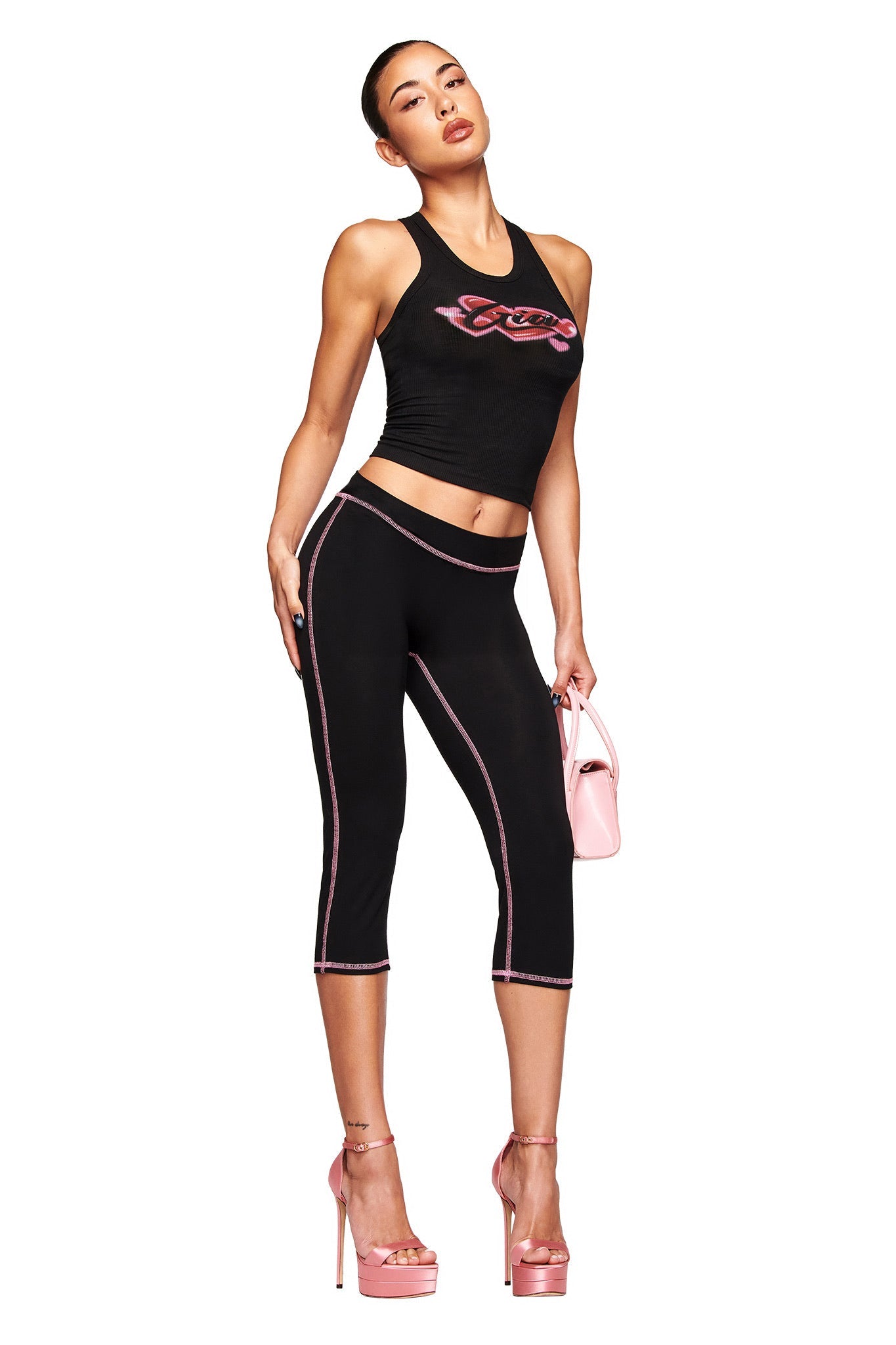 A woman models the I.AM.GIA PENELOPE TANK - GIA AIRBRUSH in black with a pink graphic, paired with black capri leggings featuring pink trim, pink platform heels, and a small pink handbag against a white background.