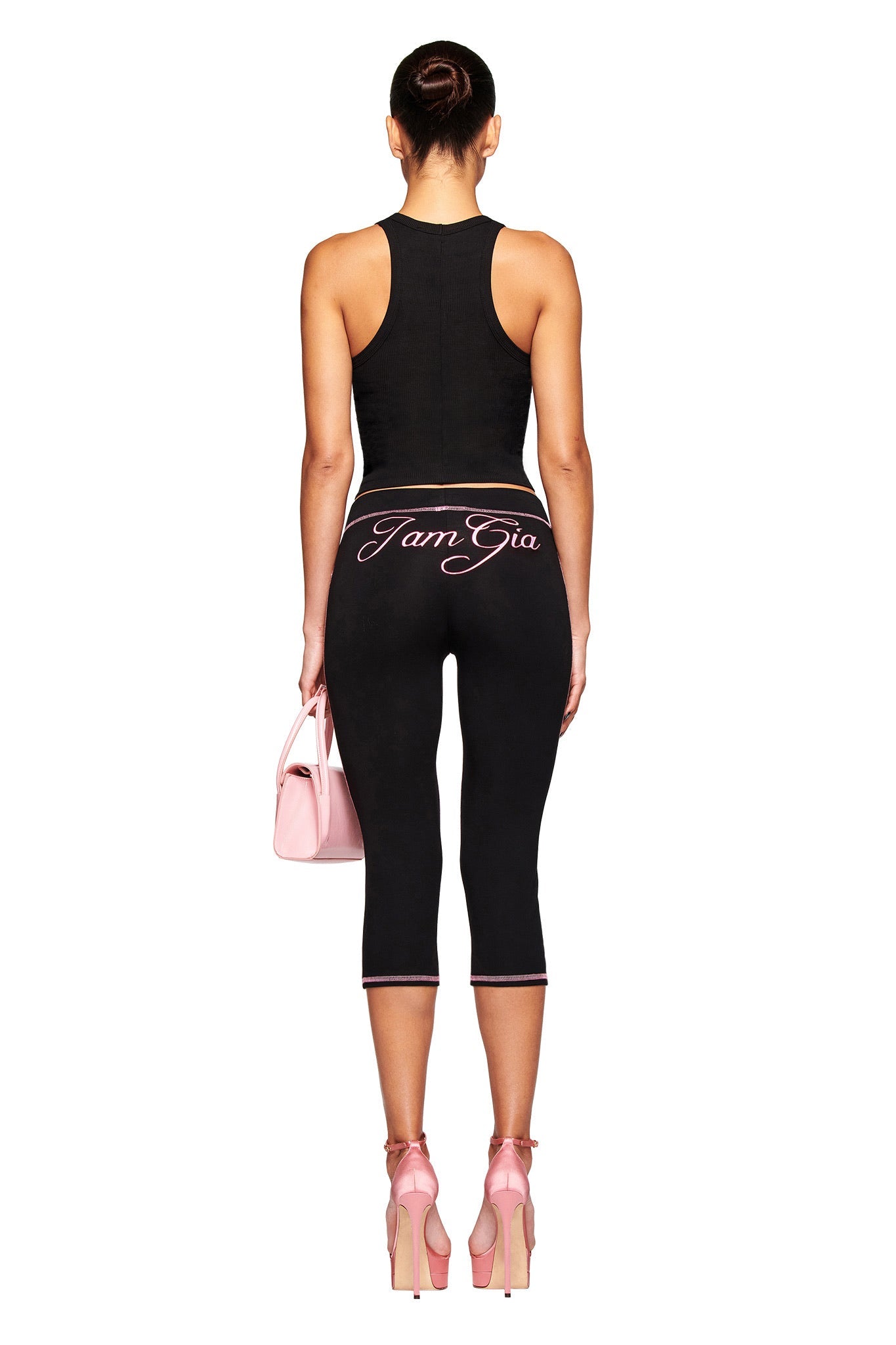 A woman faces away wearing the I.AM.GIA PENELOPE TANK - GIA AIRBRUSH and black cropped leggings with "I Am Gia" on the waistband, accessorized with a pink handbag and pink ribbon-tied high heels.