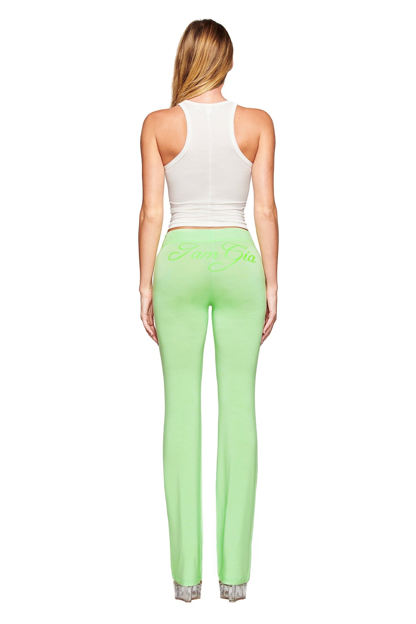 A woman with long blonde hair faces away, wearing the I.AM.GIA PENELOPE TANK - STAR AIRBRUSH in white and light green fitted pants with "Jade Gia" print on the back, posed against a white background.