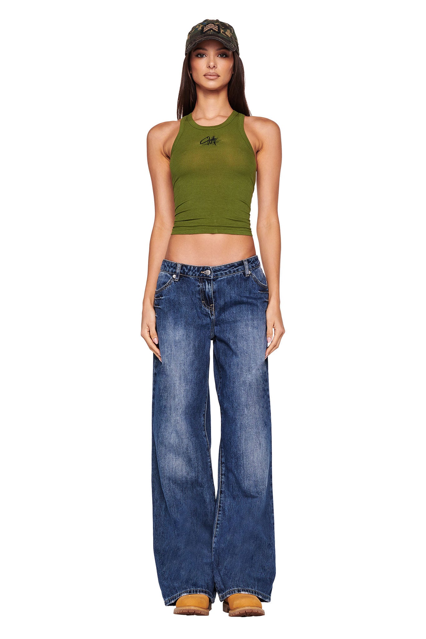 A person wearing a green crop top with a small I.AM.GIA graphic on the chest, the I.AM.GIA DYLAN JEAN in dark blue, a camo-patterned cap, and brown shoes stands facing the camera against a plain white background.