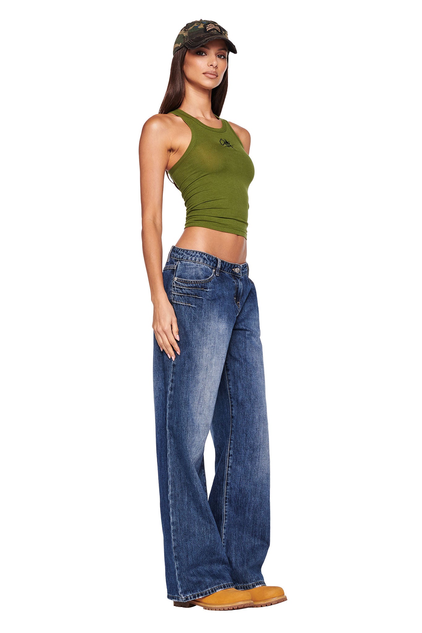 A woman stands facing slightly left, wearing a green sleeveless top by I.AM.GIA, DYLAN JEAN - BLUE : DARK BLUE wide-leg jeans, yellow work boots, and a camouflage baseball cap. She has long, straight hair and is posing confidently against a white background.