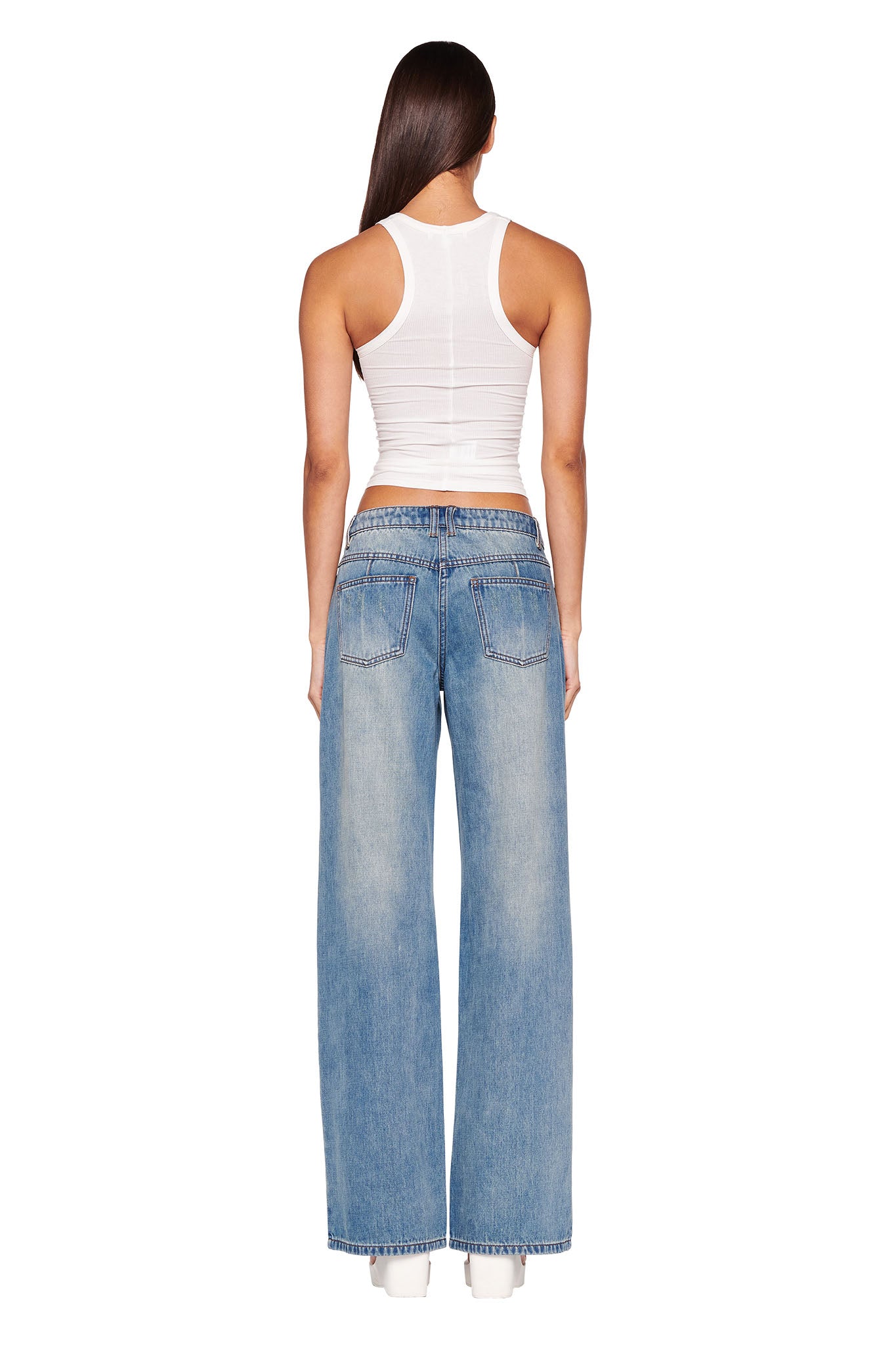 A person with long brown hair is standing with their back to the camera, wearing an I.AM.GIA white ribbed tank top and DYLAN JEAN - BLUE : LIGHT BLUE from I.AM.GIA. They are also sporting matching white shoes against a plain white background.