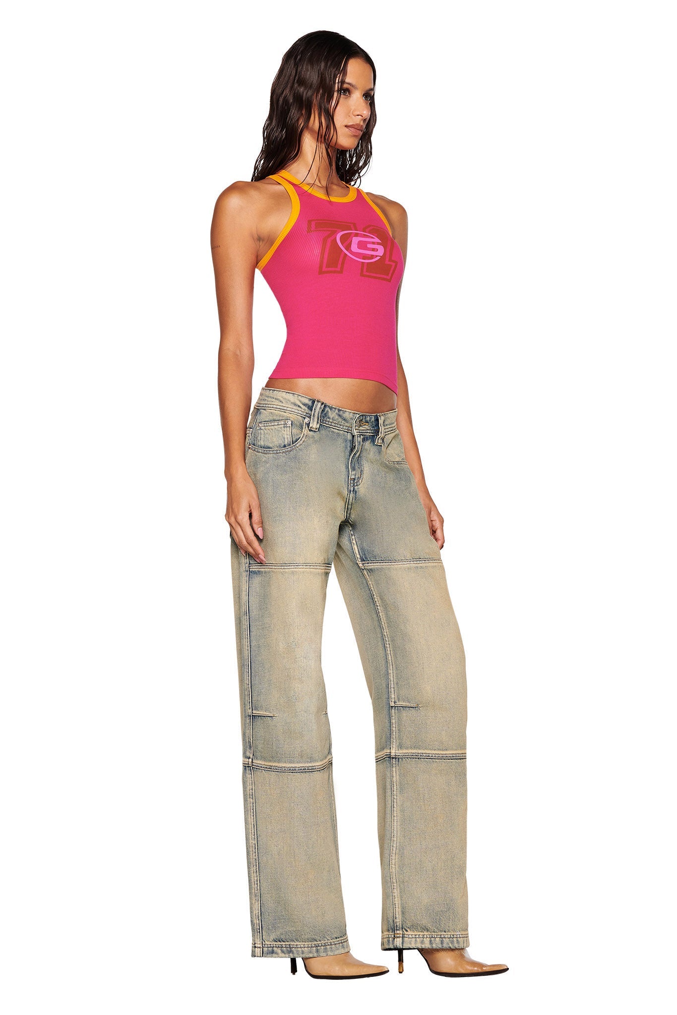 A woman stands wearing a hot pink PENELOPE TANK from I.AM.GIA, its modern design complementing the wide-leg light denim jeans. Her long dark hair cascades as she poses in beige high-heeled shoes against a plain white backdrop.