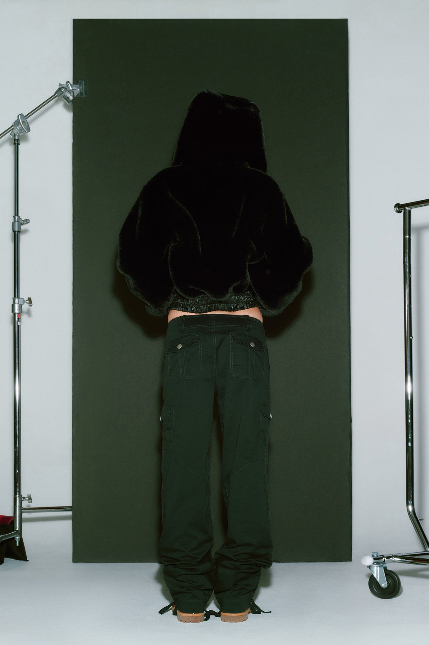 A person stands with their back to the camera, wearing an I.AM.GIA 717 Jacket in black and black pants. The individual is positioned against a black backdrop with metal stands on either side, possibly part of a photography setup.