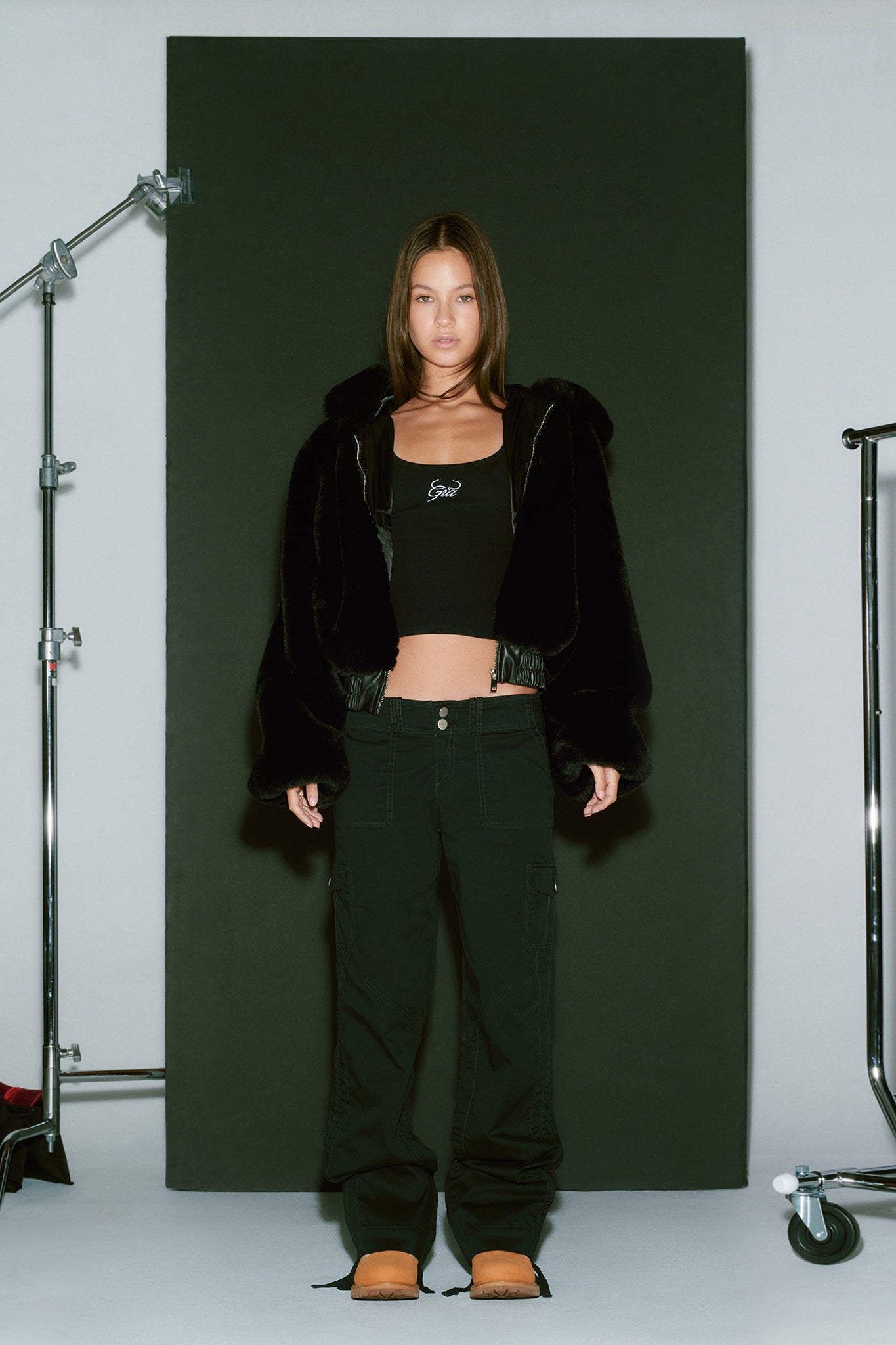 A person stands in front of a black backdrop, wearing the I.AM.GIA 717 JACKET - BLACK, a black crop top with white text, black cargo pants, and tan shoes. Metal equipment stands are visible on either side of the backdrop. The person has long, straight light brown hair.