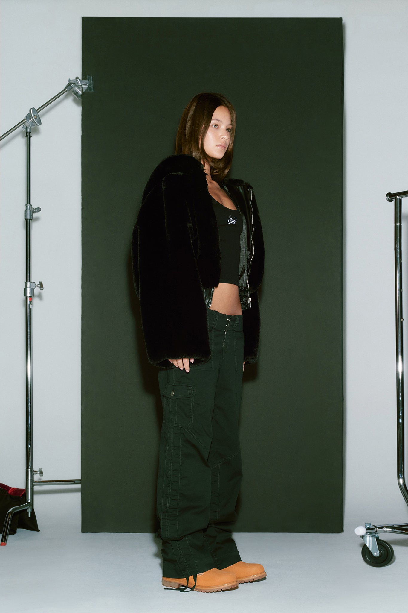 A person stands against a dark backdrop, showcasing an edgy I.AM.GIA ensemble featuring the 717 JACKET - BLACK, a black crop top, dark cargo pants, and tan work boots. Studio equipment frames the scene, with a light gray floor and off-white backdrop adding to the minimalist setting.