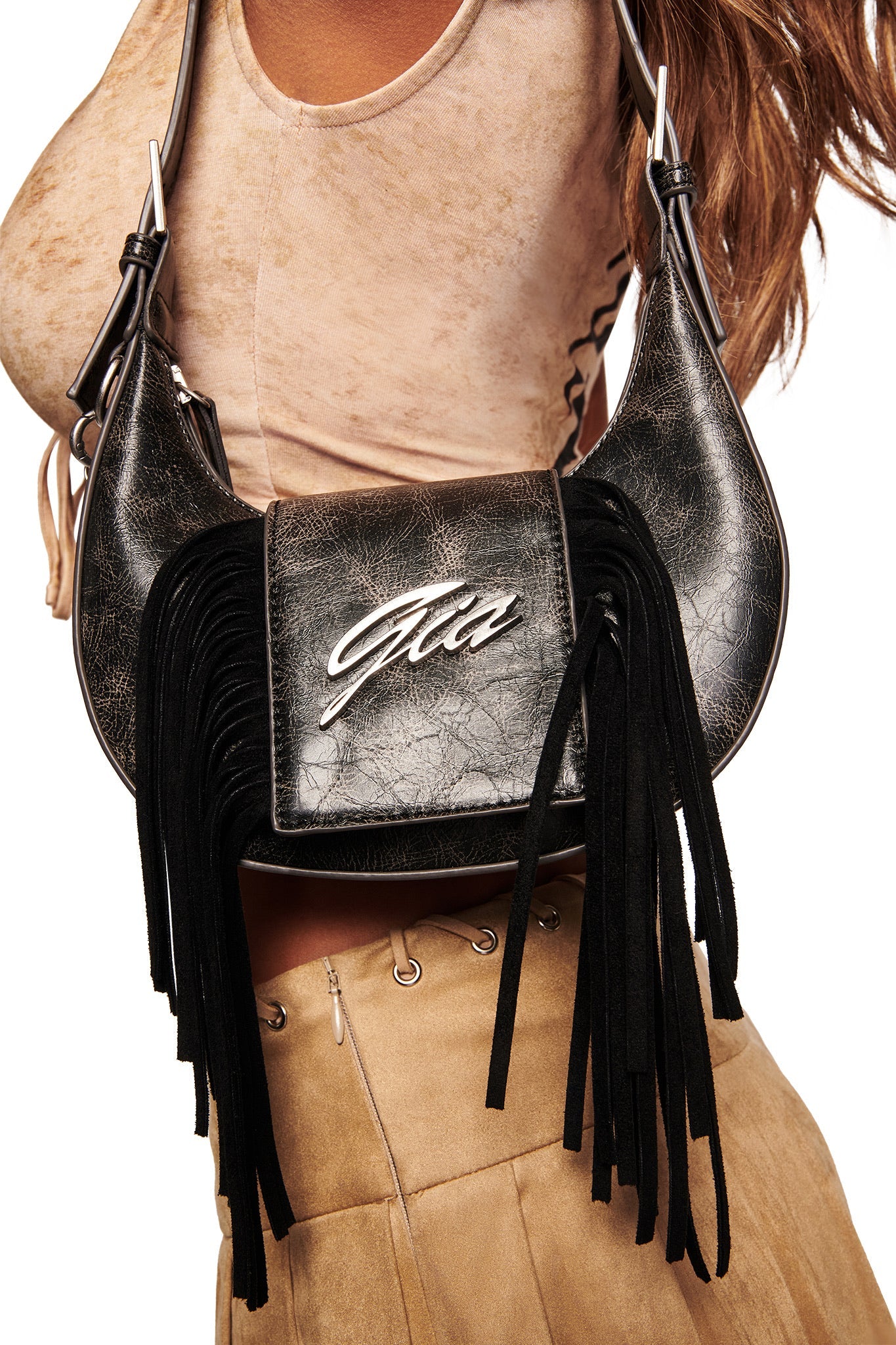 A woman in a beige crop top and tan skirt carries the I.AM.GIA TIA BAG - BLACK FRINGE, featuring black faux leather, fringe details, an adjustable strap, and a shiny silver "Gia" logo stylishly across her torso.