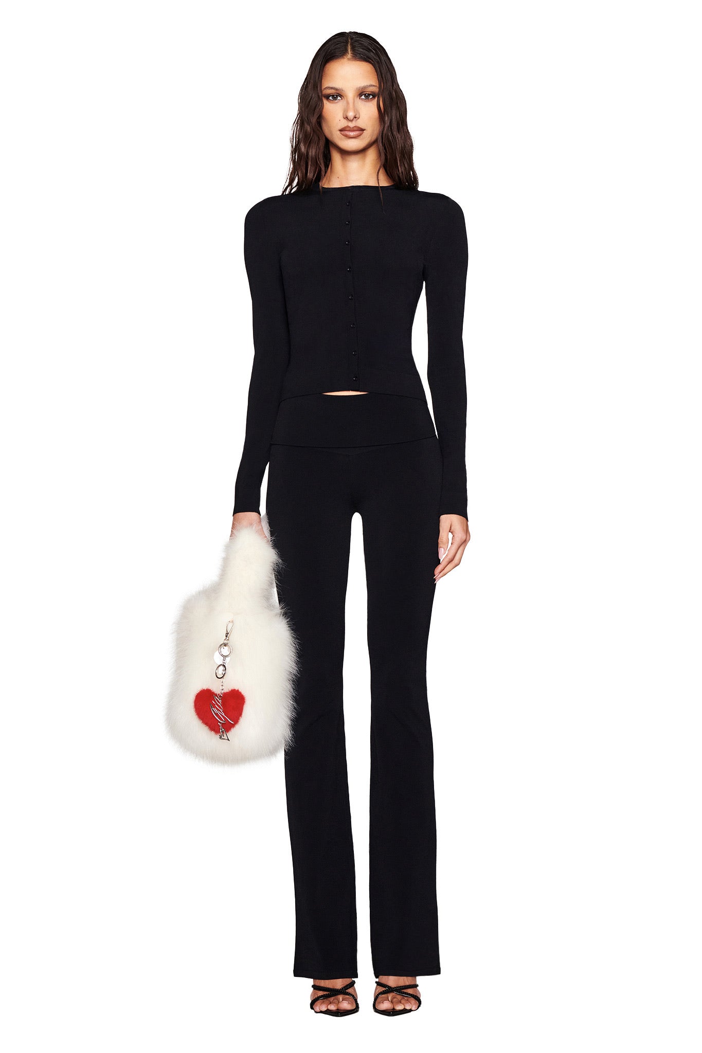A person in a fitted black long-sleeve outfit with flared pants holds the I.AM.GIA heart-shaped faux fur handbag, VALENTINA KEYCHAIN - RED, featuring a red heart and keychain detail against a plain white background.