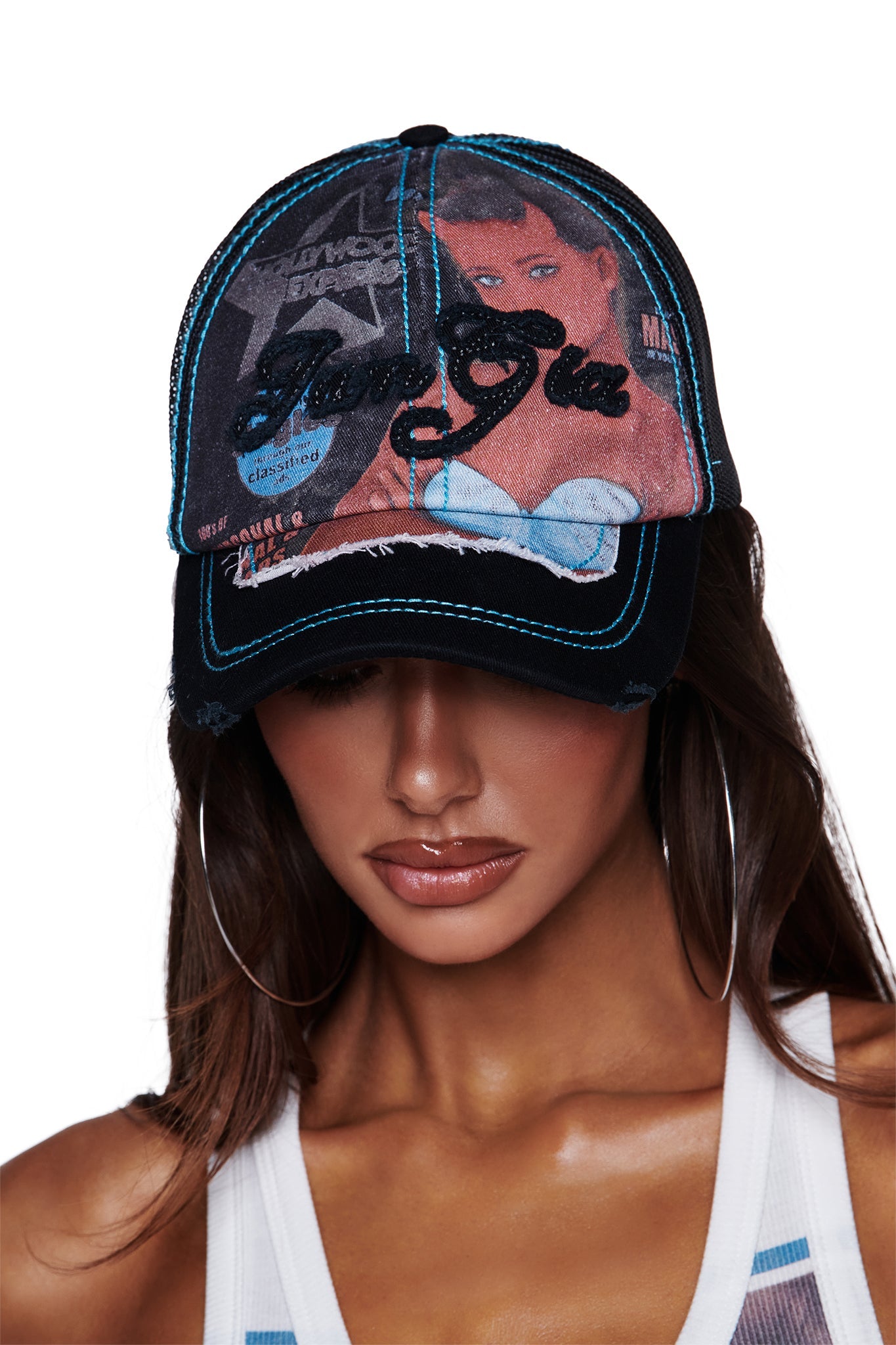 A woman wearing large hoop earrings and a tank top looks down, partially obscured by the T-REX HAT - BLACK from I.AM.GIA—a distressed black trucker hat featuring a graphic of a woman and bold I.AM.GIA artwork. The cap showcases blue stitching, a collage-style design, and an adjustable snap closure for a custom fit.