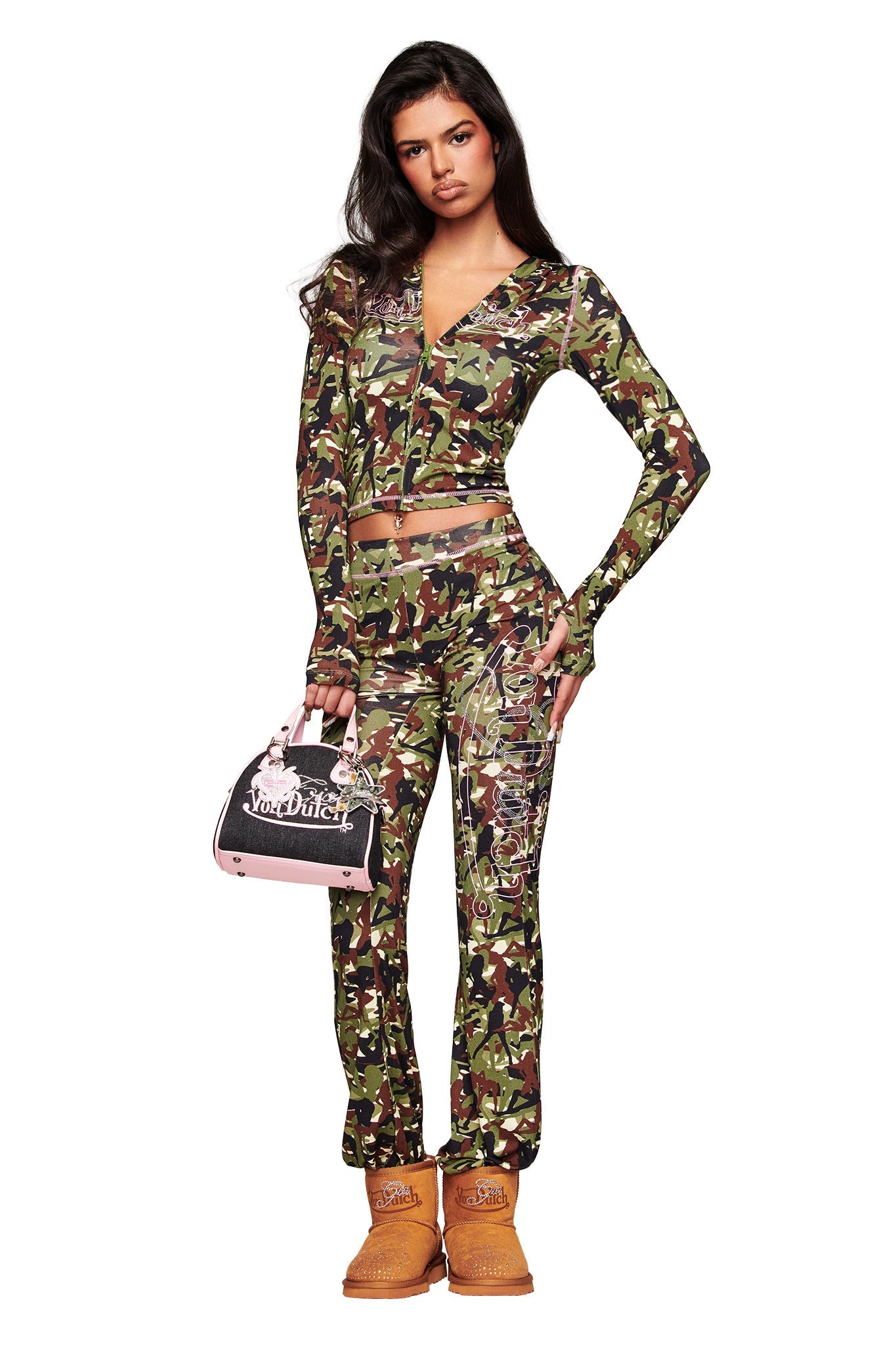 A woman with long dark hair faces forward wearing a green camo zip-up top, matching lace-up pants, tan Timberland-style boots, and holding the I.AM.GIA X VON DUTCH BOWLING BAG in indigo denim by I.AM.GIA. She has a neutral expression.