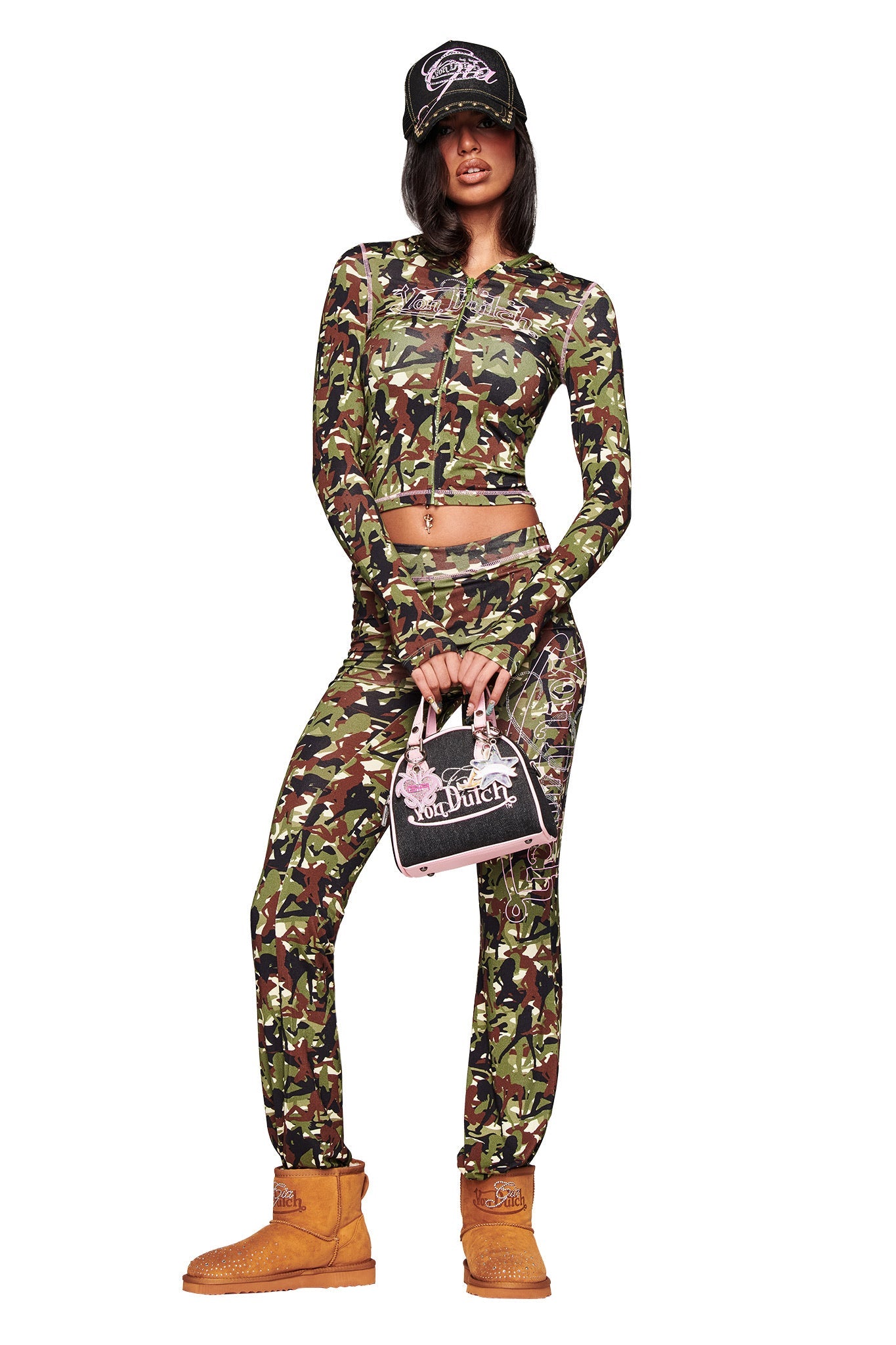 A woman poses in a green camouflage crop top and pants, black trucker hat, and carries a pink and black Von Dutch purse, completing her look with the I.AM.GIA X VON DUTCH FAUX SHEARLING BOOT in brown.