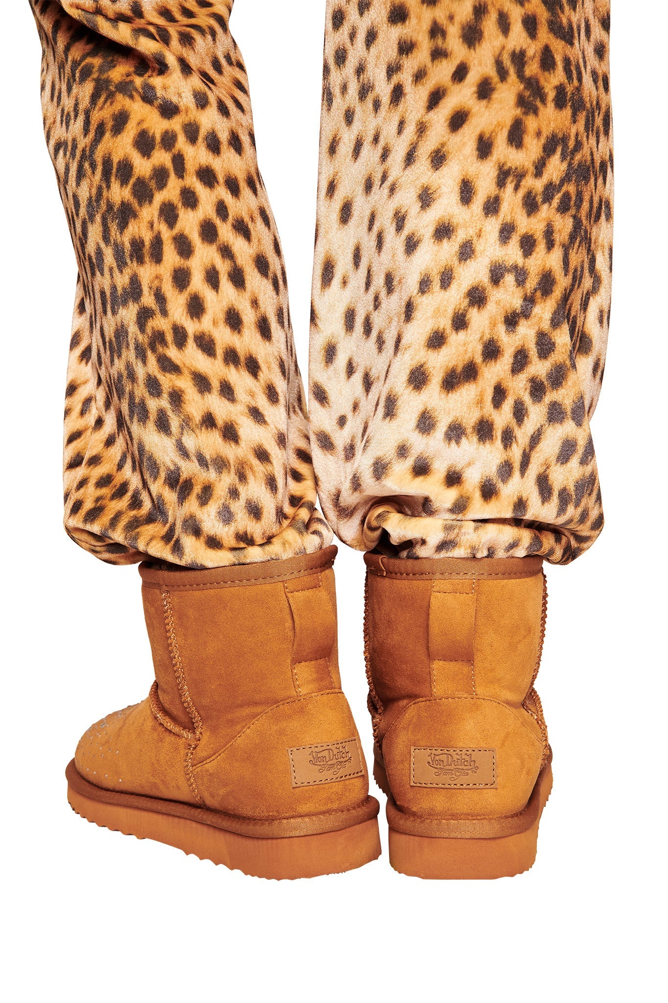 Two feet in I.AM.GIA X VON DUTCH FAUX SHEARLING BOOT - BROWN stand on a white background, paired with bold animal-print pants for an eye-catching I.AM.GIA look.