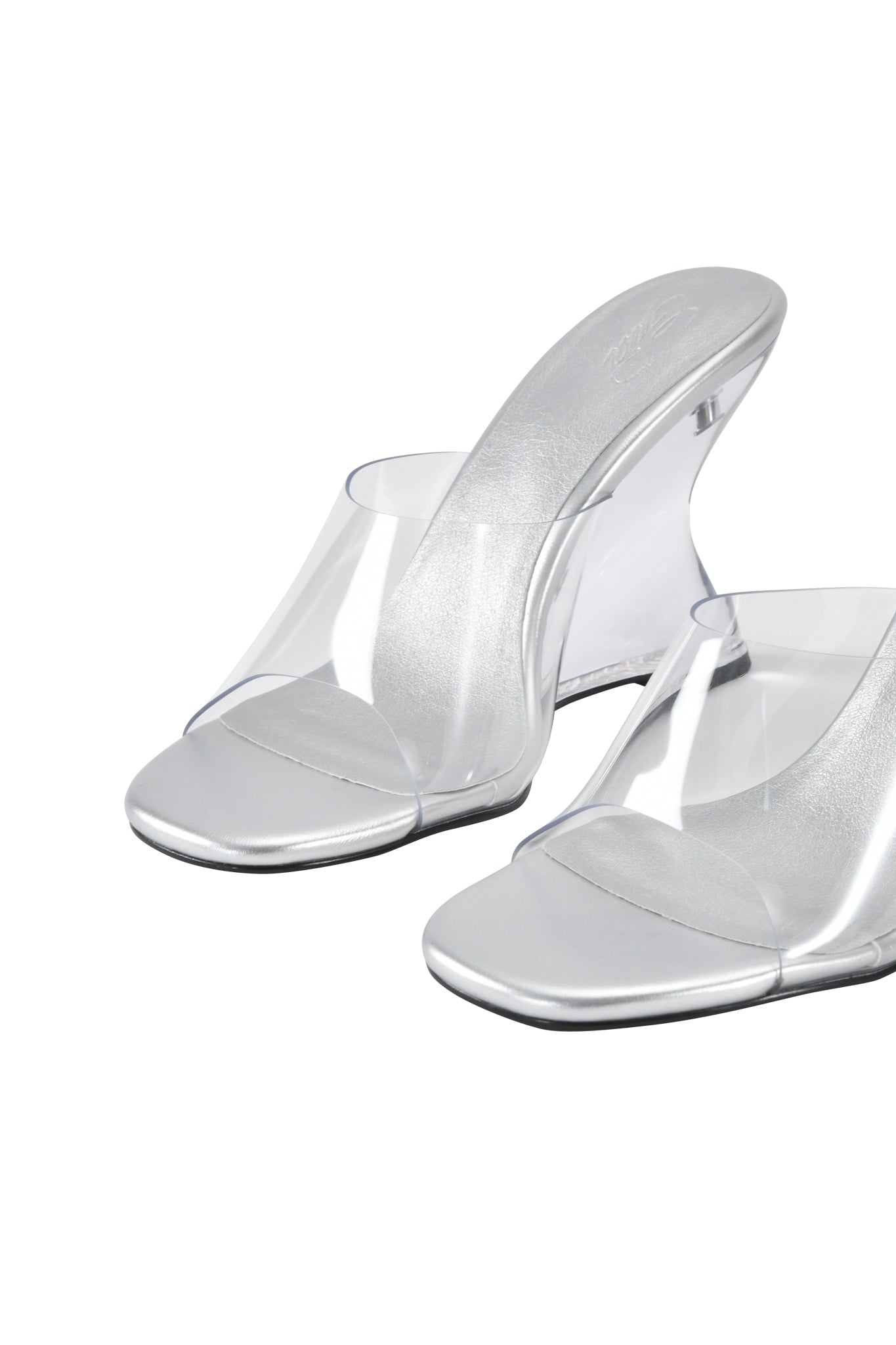 The I.AM.GIA RHEA Clear Wedge Heel - Transparent features open toes, a chic mule silhouette, clear vinyl straps, a shiny metallic footbed, and wedge heels, shown on a white background.