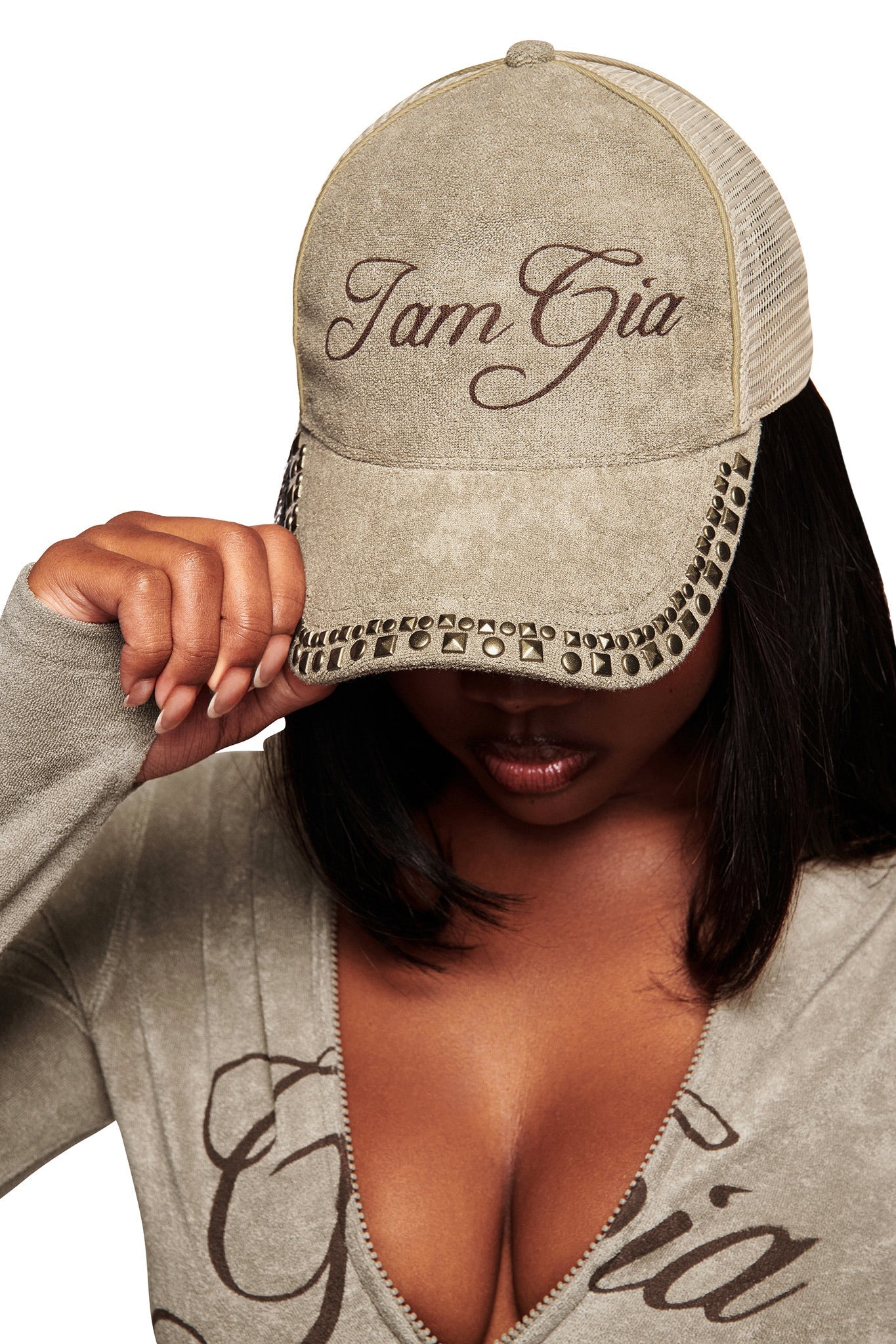 A woman with medium skin tone wears the ISSHA TERRY HAT - STONE by I.AM.GIA, a tan snapback trucker style. She pairs it with a matching low-cut top and tilts her head down, partially covering her face. White background.