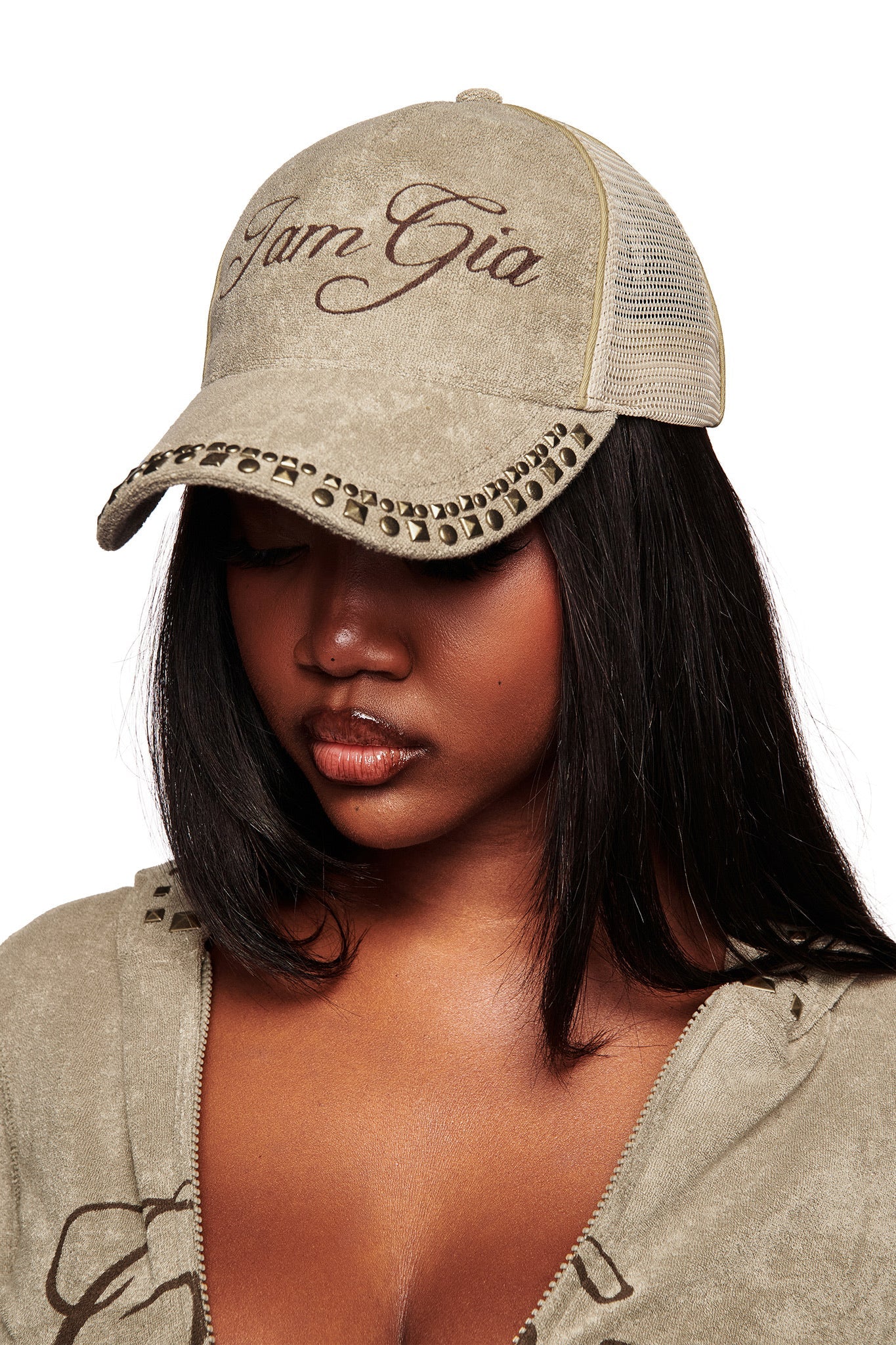 A woman with straight dark hair wears the I.AM.GIA ISSHA TERRY HAT in Stone, featuring silver studs and “IamGia” embroidery, paired with a zipped beige top against a white background.