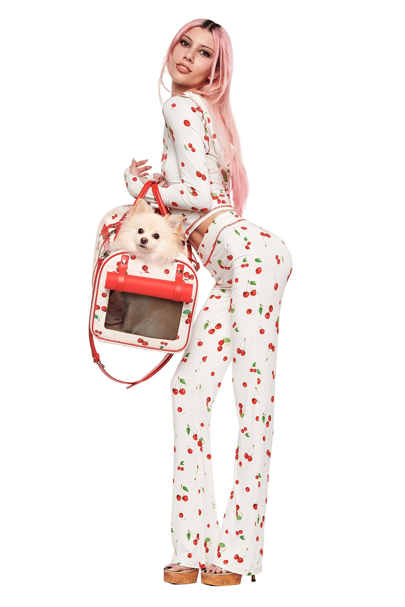 A woman with long pink hair wears a white top, flared cherry-print pants, and platform shoes. She holds her dog in the I.AM.GIA TRIXX PET CARRIER - CHERRY ON TOP and looks over her shoulder.