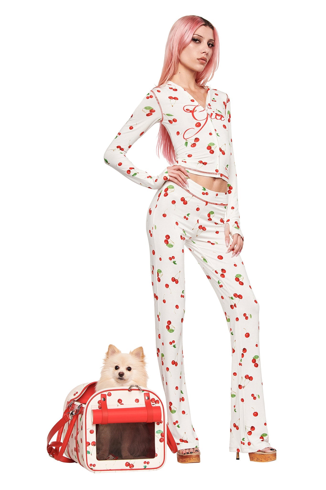 A person with long pink hair wears I.AM.GIA cherry-patterned pajamas, standing confidently beside a small fluffy dog in the matching TRIXX PET CARRIER - CHERRY ON TOP by I.AM.GIA.