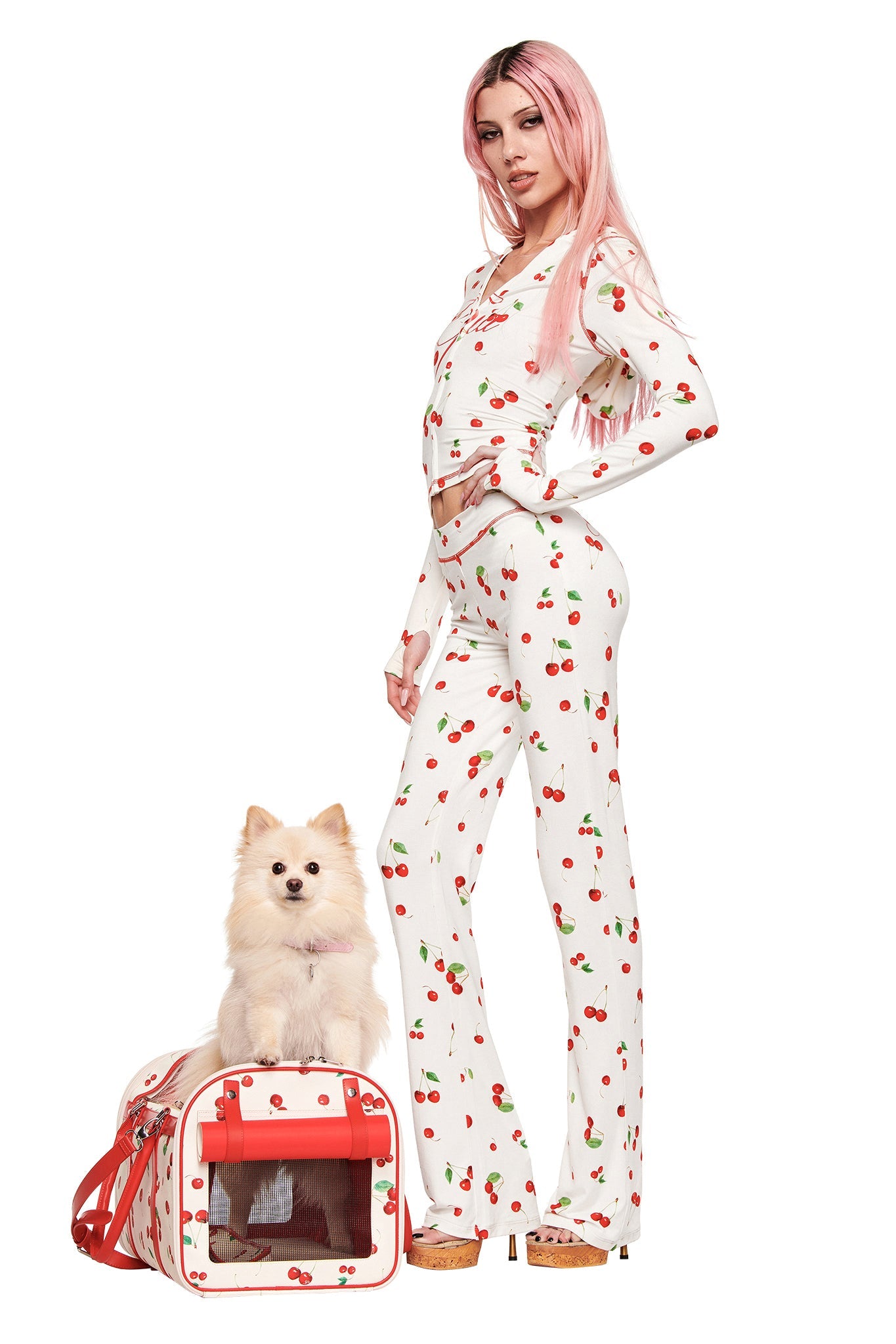 A woman with long pink hair, dressed in white cherry-print pajamas, stands with a small dog beside the I.AM.GIA TRIXX PET CARRIER – CHERRY ON TOP, a chic cherry-print small pet carrier. The background is white.