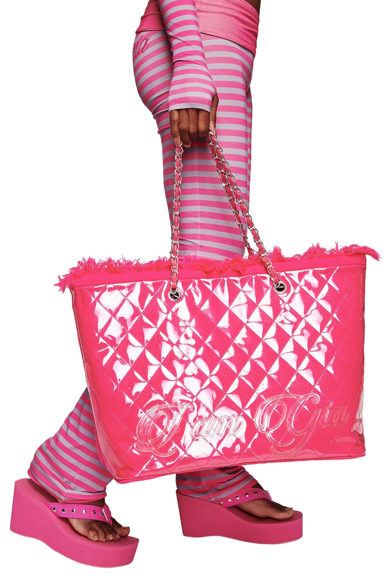 A person with pink and gray striped pants and platform sandals holds the I.AM.GIA DANNI TOTE BAG - HOT PINK, featuring chain handles and frayed edges.
