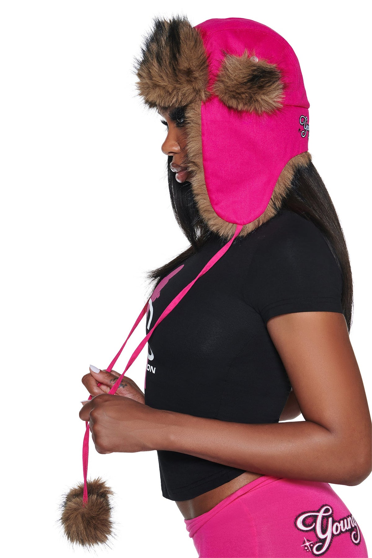 Side profile of a person wearing a bright pink YVL FUR TRAPPER HAT - PINK with brown trim and dangling pom-poms.
