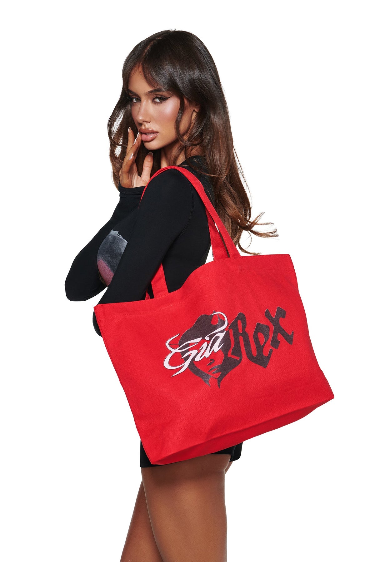 A woman in black holds the T-REX TOTE BAG - RED by I.AM.GIA, featuring bold black-and-white graphic text and devil girl artwork on a red canvas. The background is white.