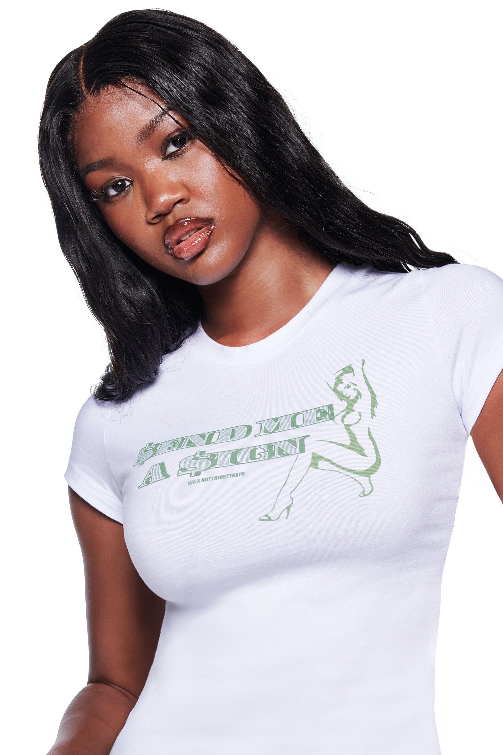 A woman with long black hair wears the I.AM.GIA POLINA DOLLA SIGN 2.0 TEE in white—a fitted cotton jersey baby tee featuring green "SEND ME A SIGN" text and a posing woman graphic—against a plain white background.