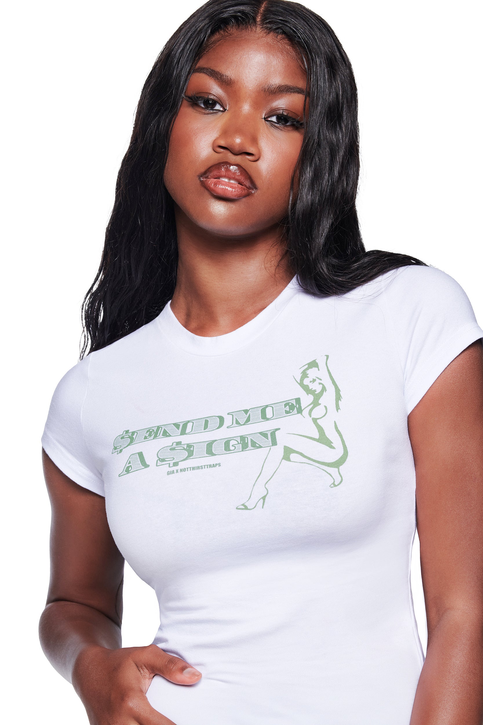 A woman with long black hair stands against a white background wearing the I.AM.GIA POLINA DOLLA SIGN 2.0 TEE in white, featuring “Send Me A Sign” in green and an outline of a seated woman.