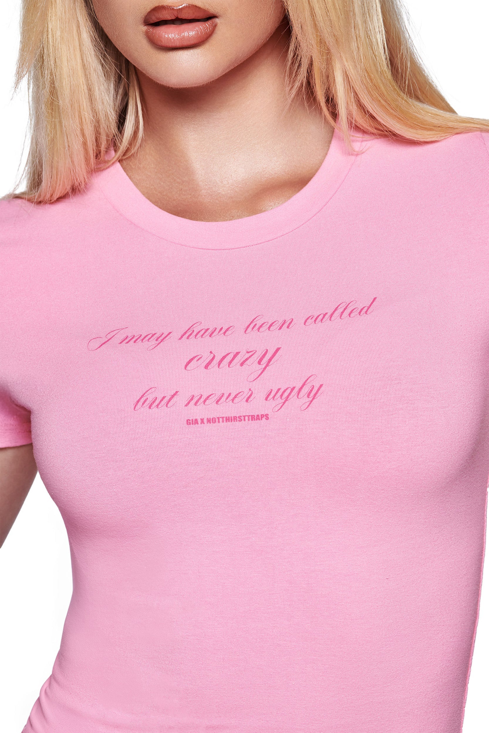 A woman wears the I.AM.GIA POLINA CRAZY TEE in pink, a cotton jersey baby tee featuring "I may have been called curvy but never ugly" printed in cursive across the chest.