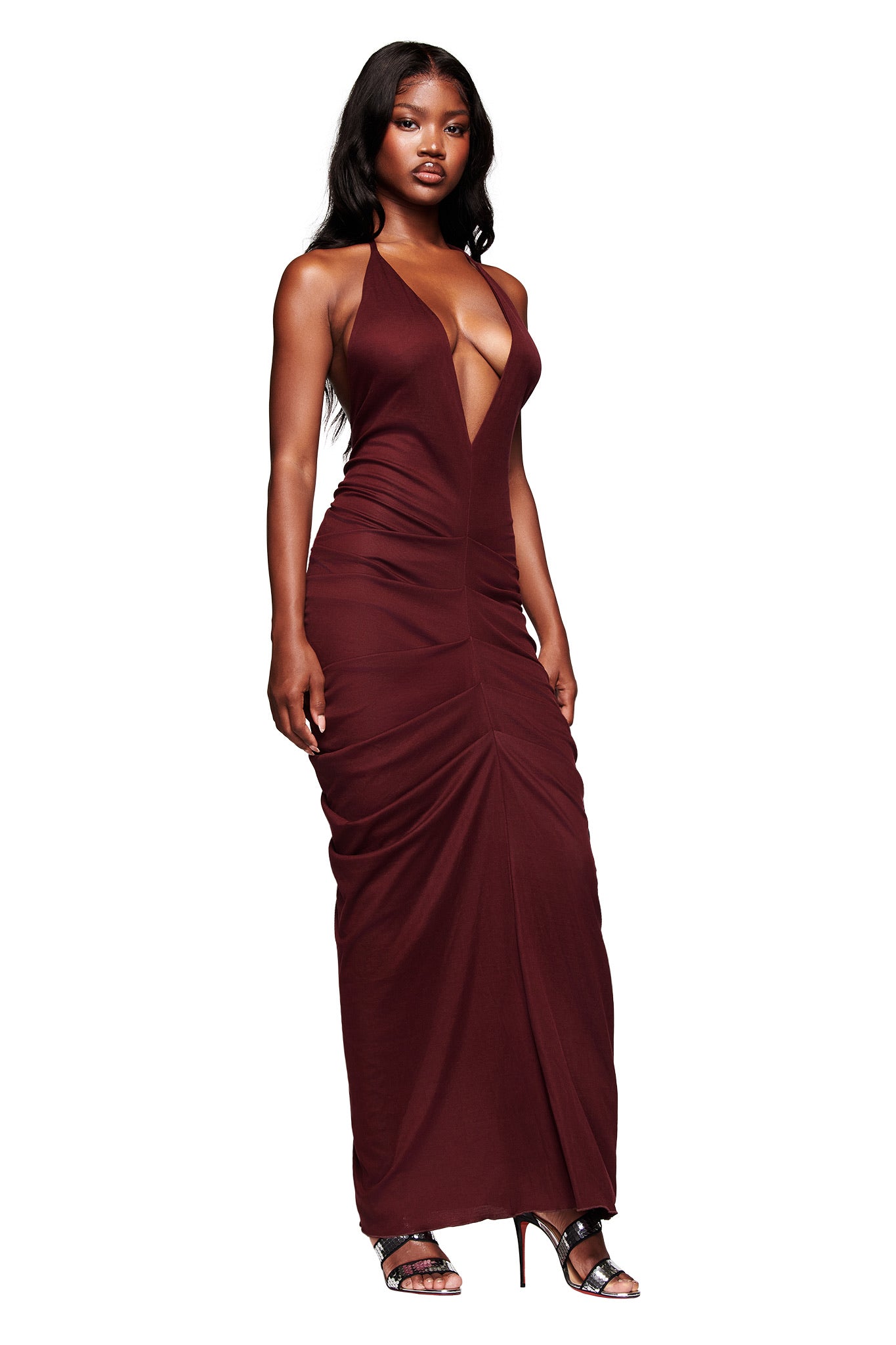 A woman in the I.AM.GIA US SASHI MAXI DRESS - PLUM, featuring a sleeveless, deep V-neck halter style with ruched detail, stands in strappy high heels against a white background.