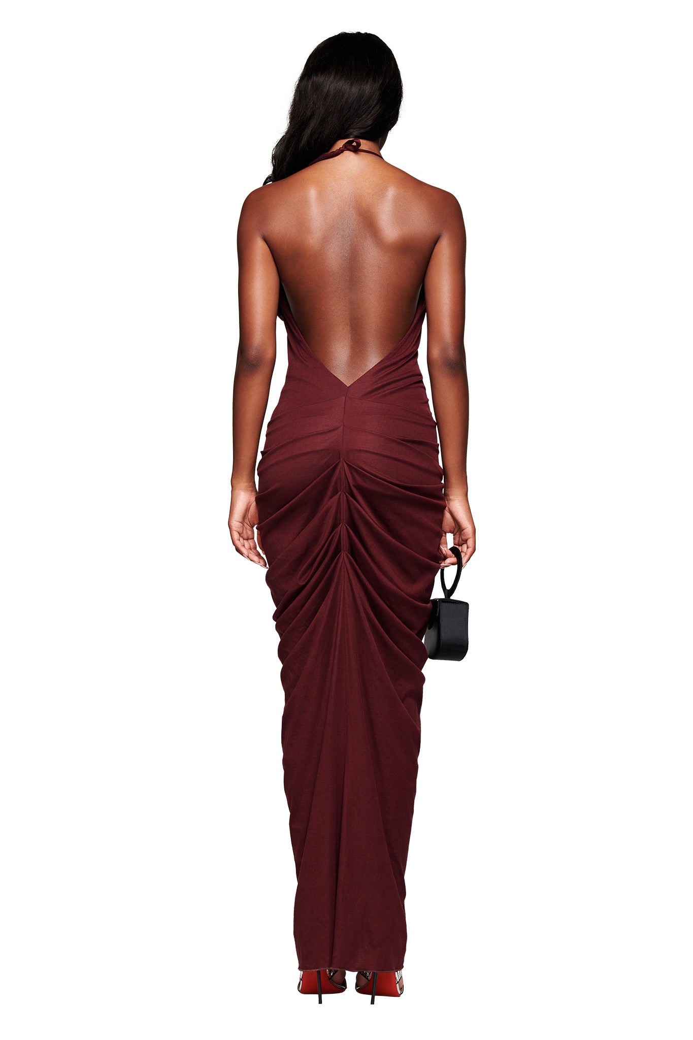 A woman with long hair wears the I.AM.GIA US SASHI MAXI DRESS in plum, featuring ruched details. She holds a small black handbag in her left hand and wears red shoes. The background is white.