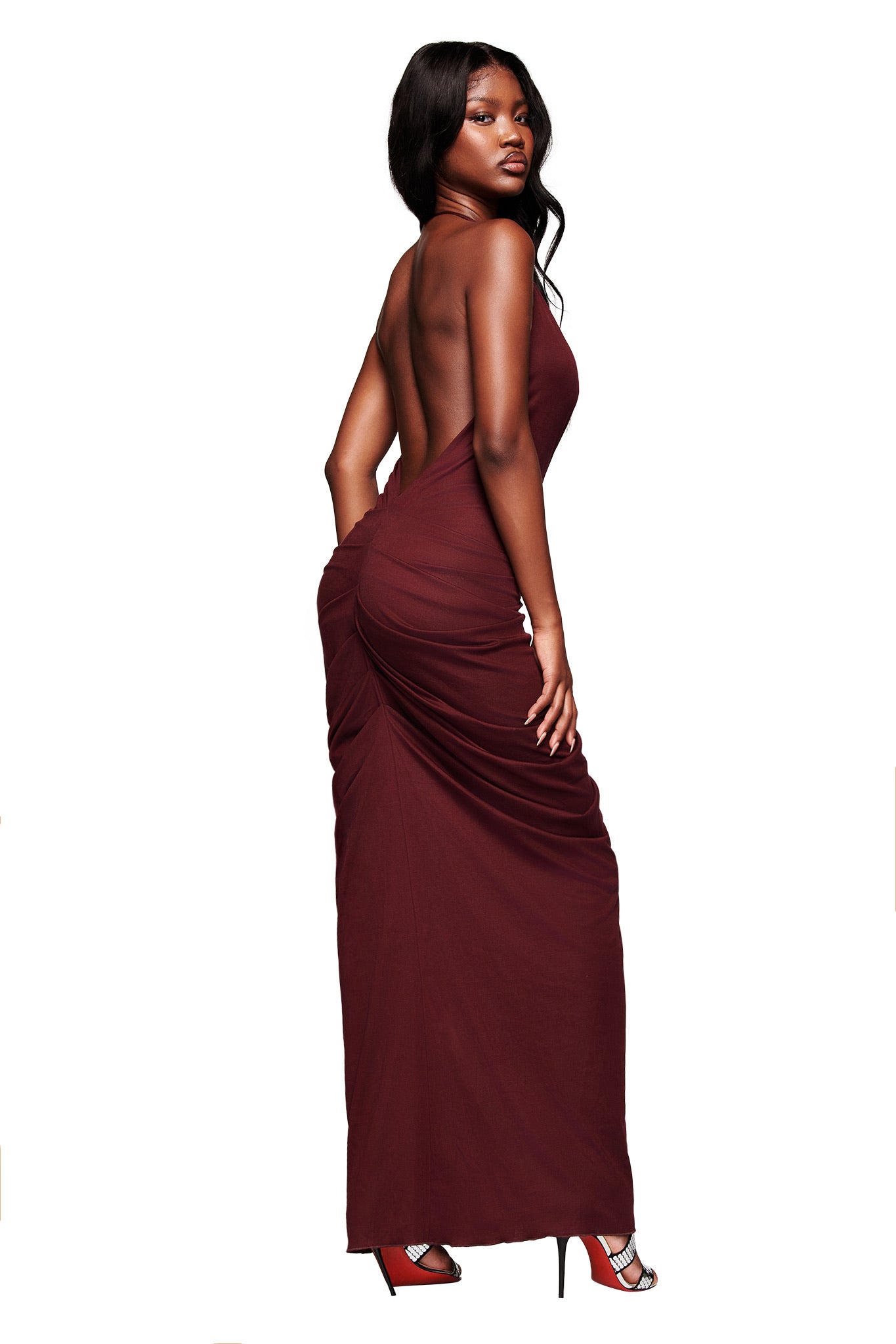 A woman models the SASHI MAXI DRESS in plum by I.AM.GIA US, a backless, form-fitting halter V-neck dress with ruched detailing, paired with red heels. She stands against a white background, looking over her shoulder.