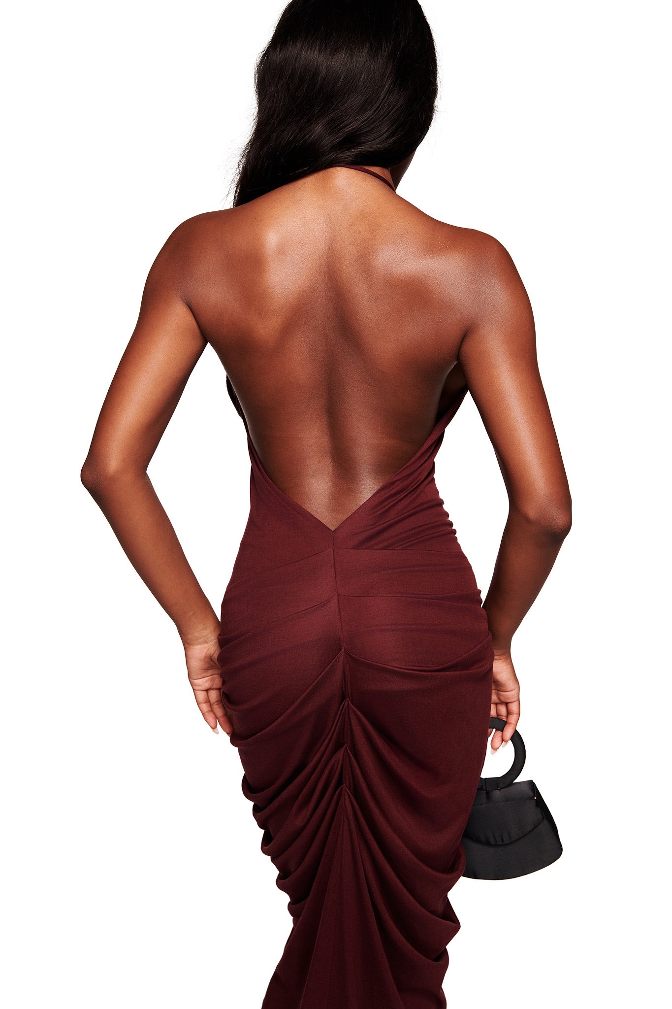 A woman with long dark hair wears the I.AM.GIA US SASHI MAXI DRESS in plum, featuring a halter V-neck and ruching detail, and holds a small black handbag against a white background.