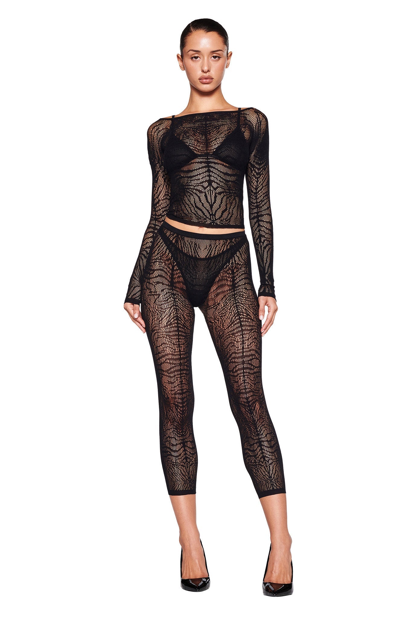 A model stands against a plain backdrop wearing the SYBIL CAPRI - BLACK : ZEBRA bodysuit from I.AM.GIA, which boasts an intricate, web-like pattern reminiscent of a netted stocking mesh. The bodysuit features long sleeves, a round neckline, and ankle-length legs. Black high-heeled shoes complete the outfit.