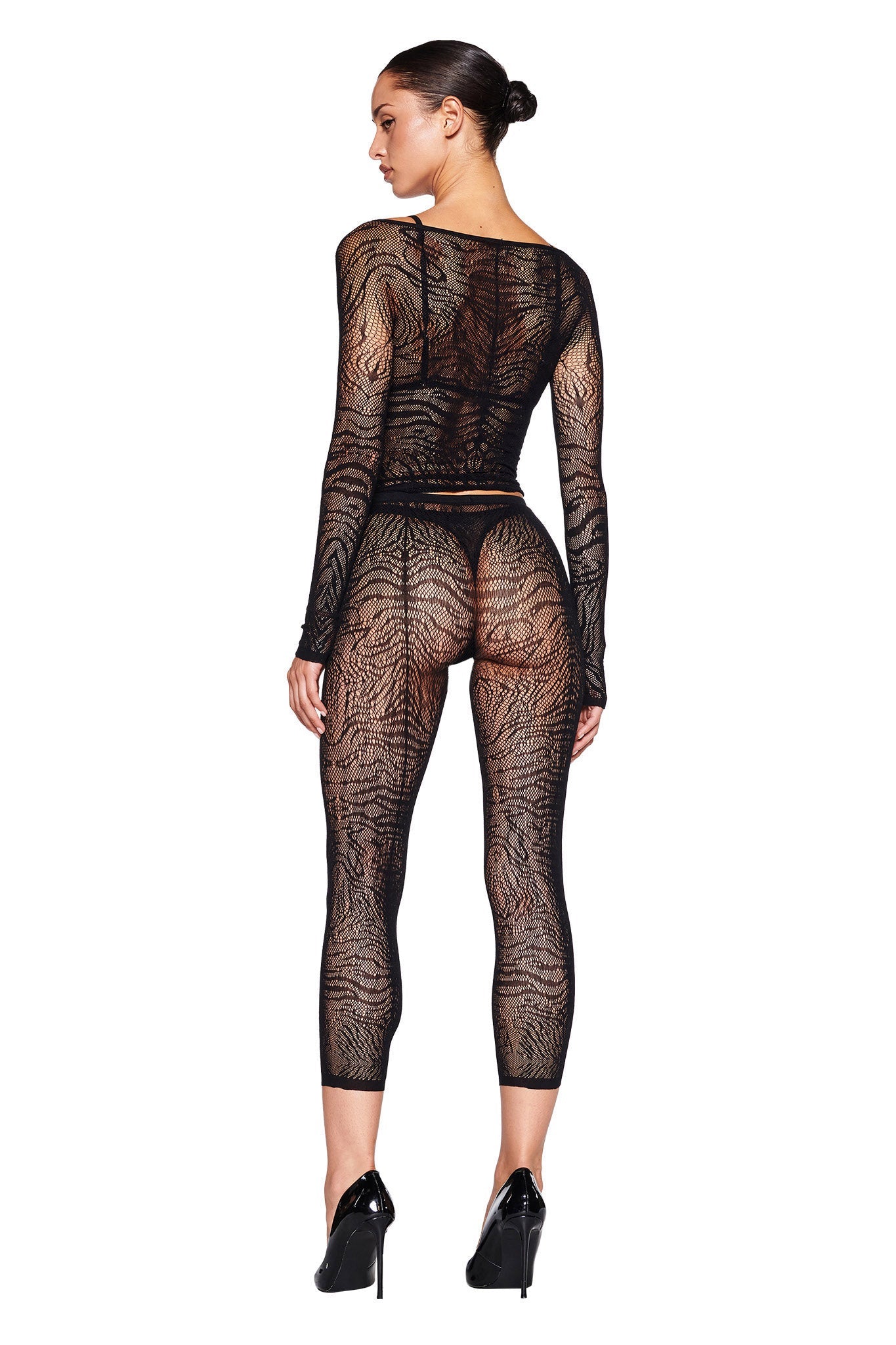 A woman is facing away, wearing a sheer black long-sleeved bodysuit with a tiger-stripe pattern and custom leopard print. Her hair is pulled back into a neat bun as she stands confidently in black high heels. The form-fitting SYBIL CAPRI - BLACK : ZEBRA by I.AM.GIA extends to her ankles, exuding elegance and edge.