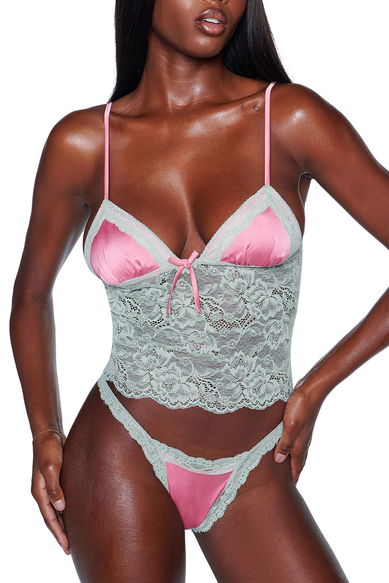 A person wearing the LOLO 2 PIECE SET - PINK by I.AM.GIA showcases a satin lace lingerie ensemble featuring a pink satin bra with green lace overlay, complemented by matching green lace bottoms. This design includes thin straps and a bow detail on the bra. The model exudes confidence while posing with one hand on their hip.