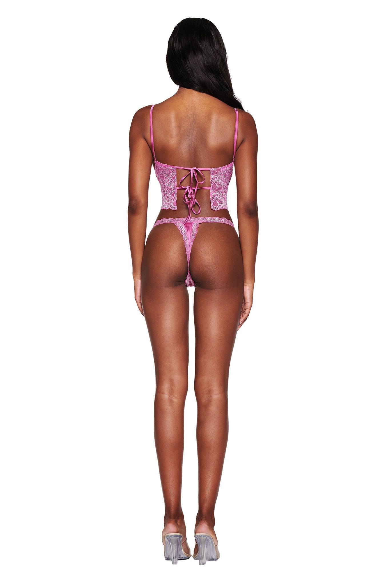 A person stands in clear high heels wearing the I.AM.GIA LOLO METALLIC SET - MAGENTA, featuring a metallic lace corset with lace-up back detail and a matching low rise thong. Long, dark hair cascades down their back.