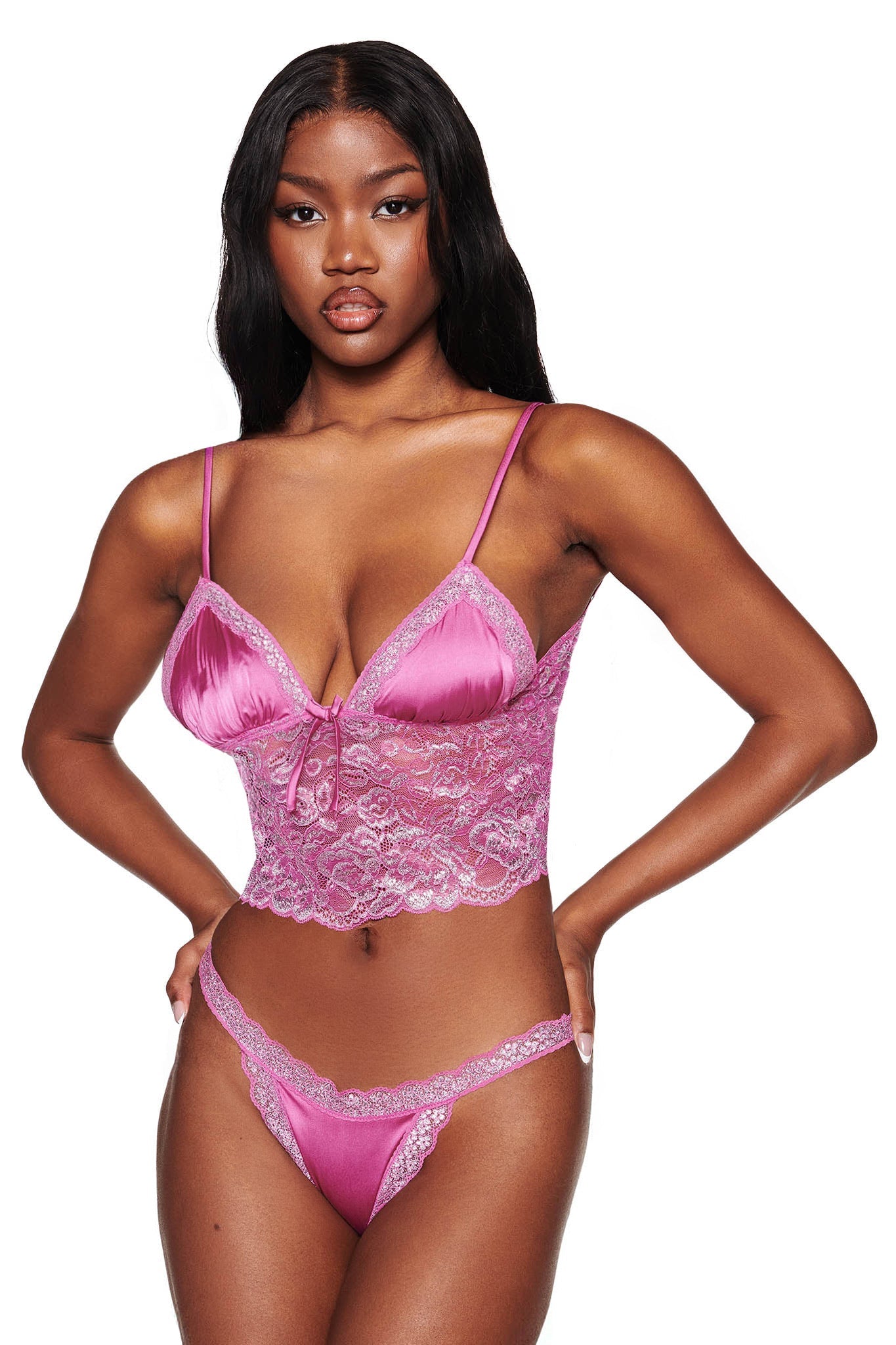 A woman confidently poses in the I.AM.GIA LOLO METALLIC SET - MAGENTA, featuring pink lace with a lace-up design and floral patterns. The set includes adjustable shoulder straps and a low-rise thong, complementing her long, dark hair against a plain white background.