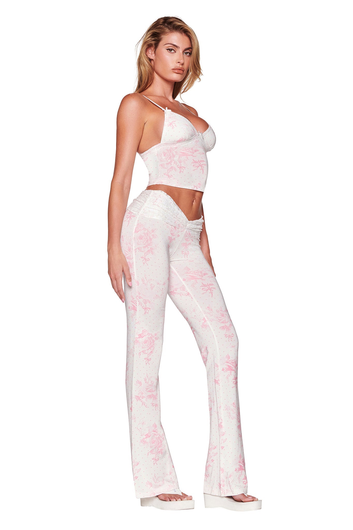 A person poses in a white cropped camisole and the I.AM.GIA KENDRA PANT in a bunny bouquet floral print, featuring a semi-sheer fabric with a low-rise flared silhouette. They complete the look with white platform shoes, set against a plain white background.