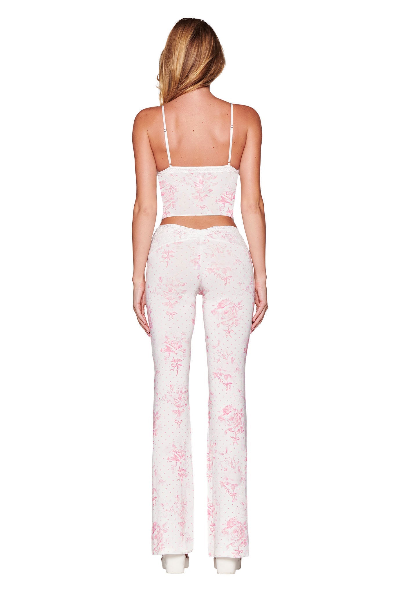 A person with long blonde hair is wearing the KENDRA TANK - WHITE: FLORAL: BUNNY BOUQUET and matching high-waisted flared pants from I.AM.GIA, featuring a delicate pink pattern. This ensemble inspired by Kendra styles stands out beautifully against the plain white background.
