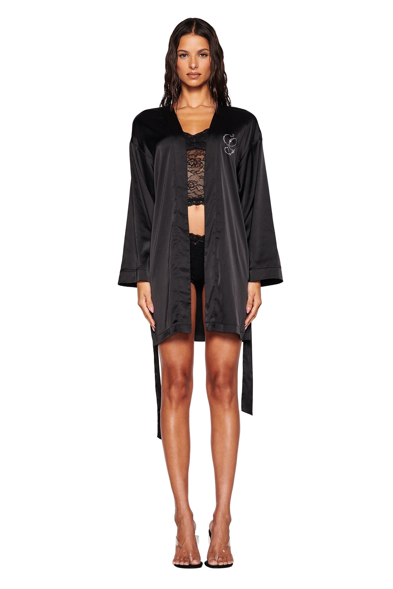 A person stands facing forward, wearing the sophisticated LARSA 2.0 SLEEP ROBE - BLACK by I.AM.GIA, crafted from sumptuous satin material over a lace top and black shorts. The robe features an intricate butterfly diamante design on the chest. They are complemented with clear, strappy high-heeled shoes against a plain white backdrop.