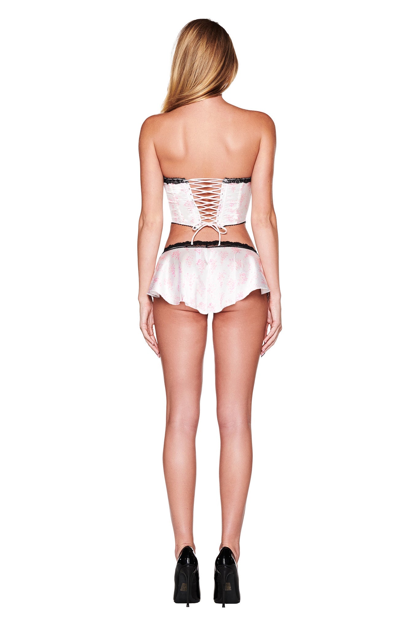 A woman with long hair faces away from the camera, displaying the BRIDGET BUSTIER 2 PIECE SET from I.AM.GIA, which features vintage-inspired delicate black lace trims. She pairs it with a frilly miniskirt and high heels. Her strapless corset outfit is primarily in a soft pink floral design complemented by tasteful black accents.