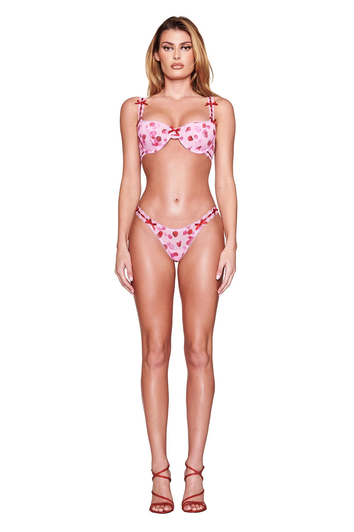 A person wears the I.AM.GIA GABRIELLA 2 PIECE SET in Pretty Petal, showcasing a pink bikini with heart patterns and red bow accents against a white background. Red high-heeled sandals complete the look.