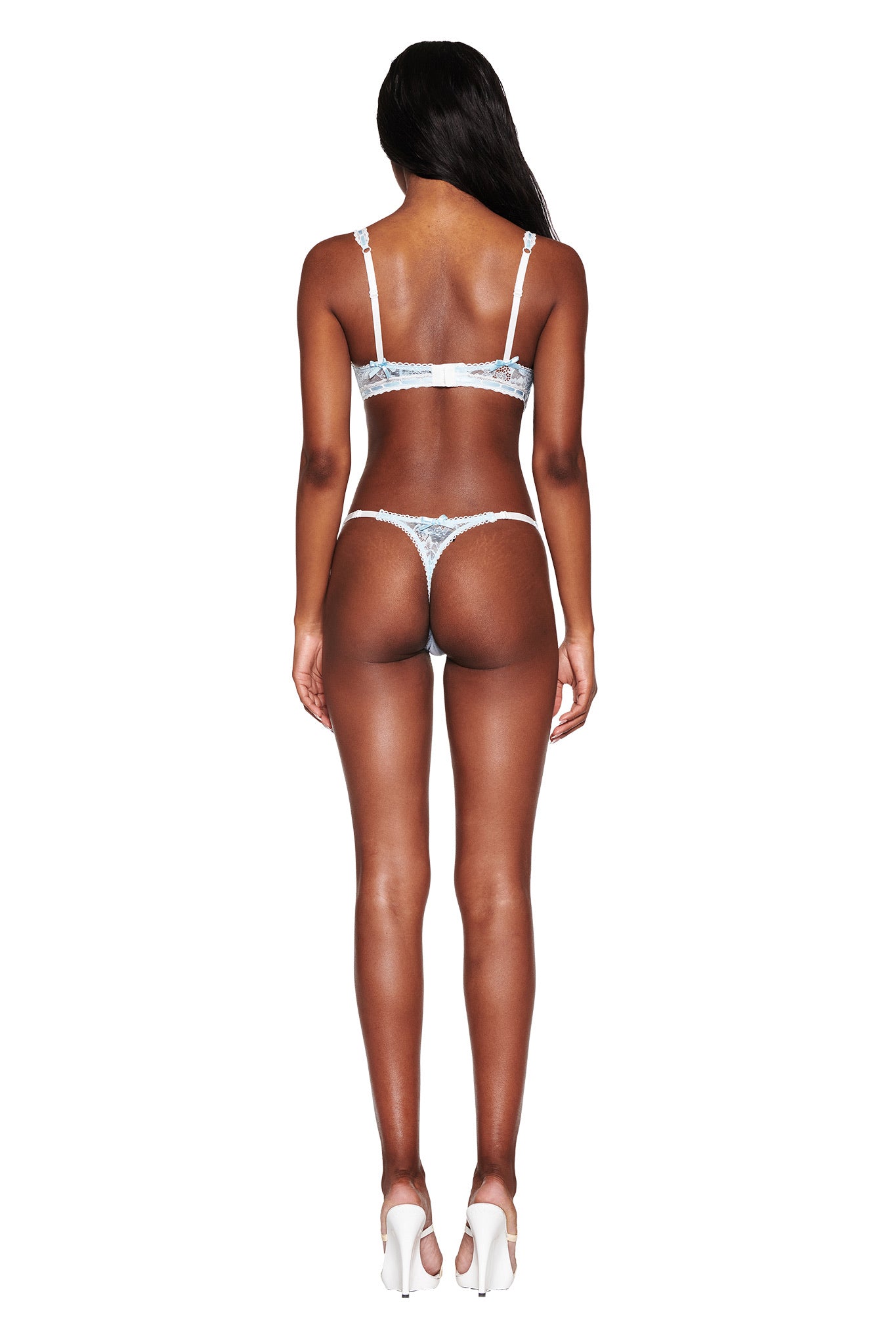 Facing away from the camera, a person models the I.AM.GIA ROMY 2 PIECE SET - BLUE, featuring an elegant bra and thong with embroidered daisy lace. Sleek in white high heels, its adjustable straps promise a flawless fit against the plain white backdrop.