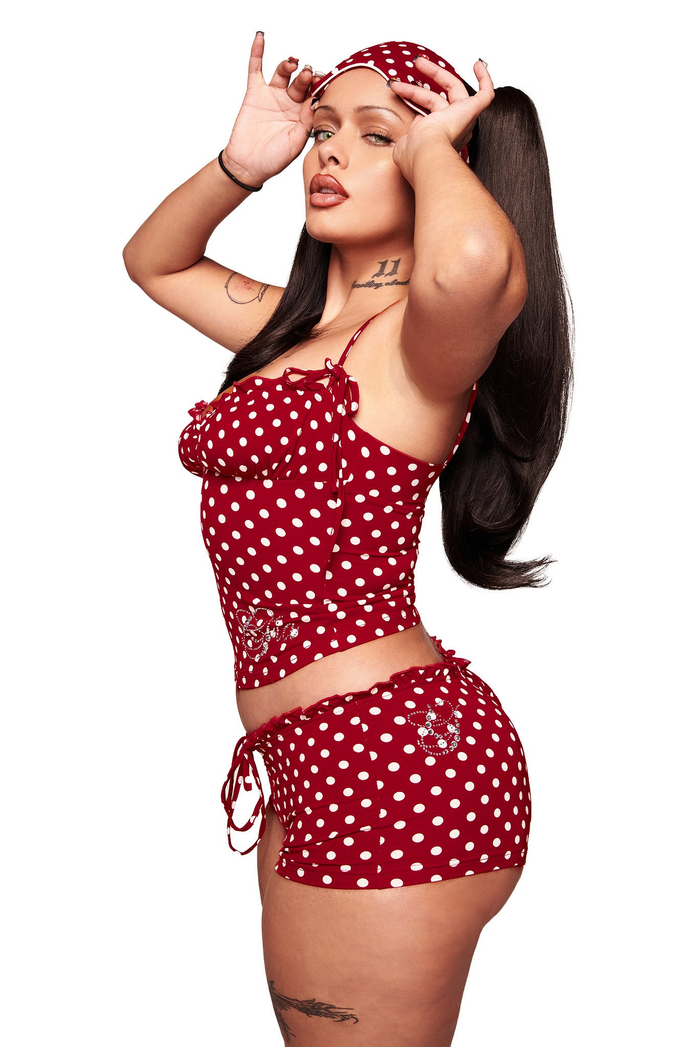 A woman poses confidently against a white background wearing the I.AM.GIA MELANIE CAMI in red polka dot with an adjustable bust fit and high-waisted shorts, her hands near her face.