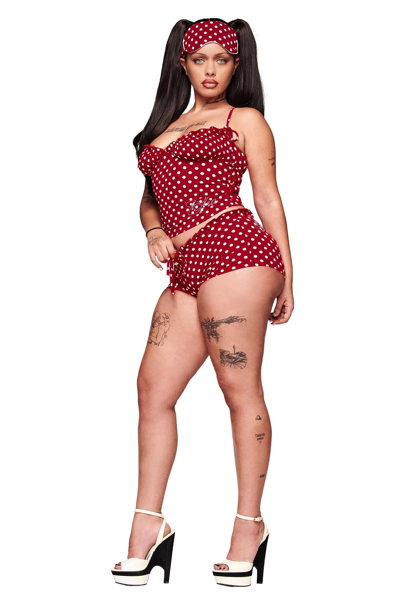 A woman models the I.AM.GIA MELANIE CAMI - RED POLKA DOT sleepwear set with adjustable bust, paired with a matching headband and white platform heels. She has visible tattoos and long dark pigtails, posed against a white background.